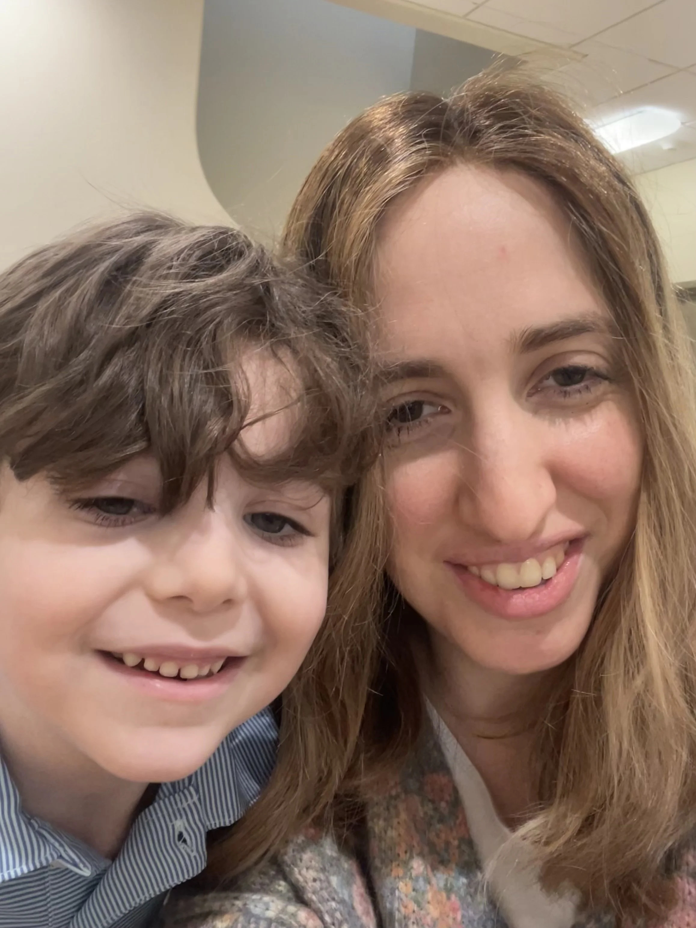 A woman with long light brown hair and a young boy with dark brown curly hair are taking a close-up selfie together, both smiling.