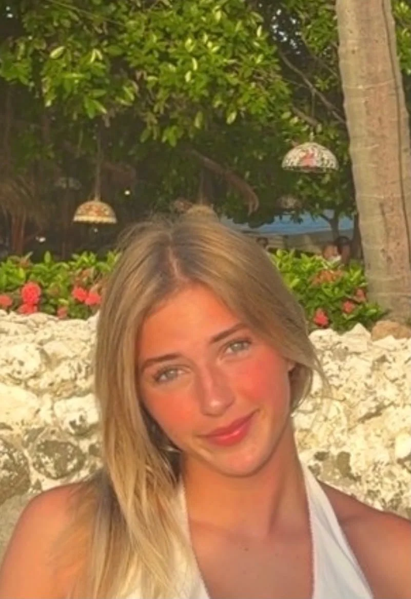 A young woman with long blonde hair smiling outdoors at a tropical location with lush greenery, pink flowers, and hanging lamps in the background.