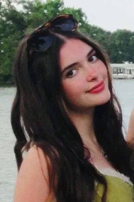 A young woman with long dark hair and sunglasses on her head, smiling outdoors near a body of water with trees and a building in the background.