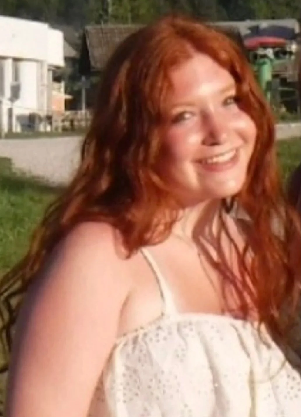 A woman with long red wavy hair smiling outdoors wearing a white summer dress.