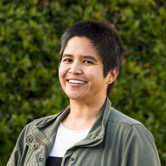 A young woman with short dark hair smiling outdoors, wearing an olive green jacket and black and white color block sweater, with greenery in the background.