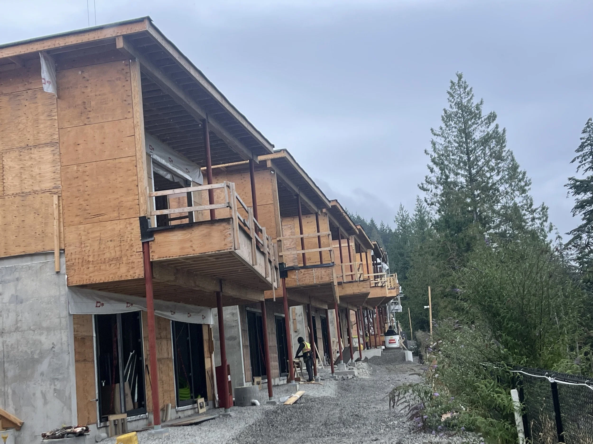 Row of multi-story buildings under construction, with unfinished wooden exteriors and scaffolding, situated in a wooded area with trees and a cloudy sky in the background.