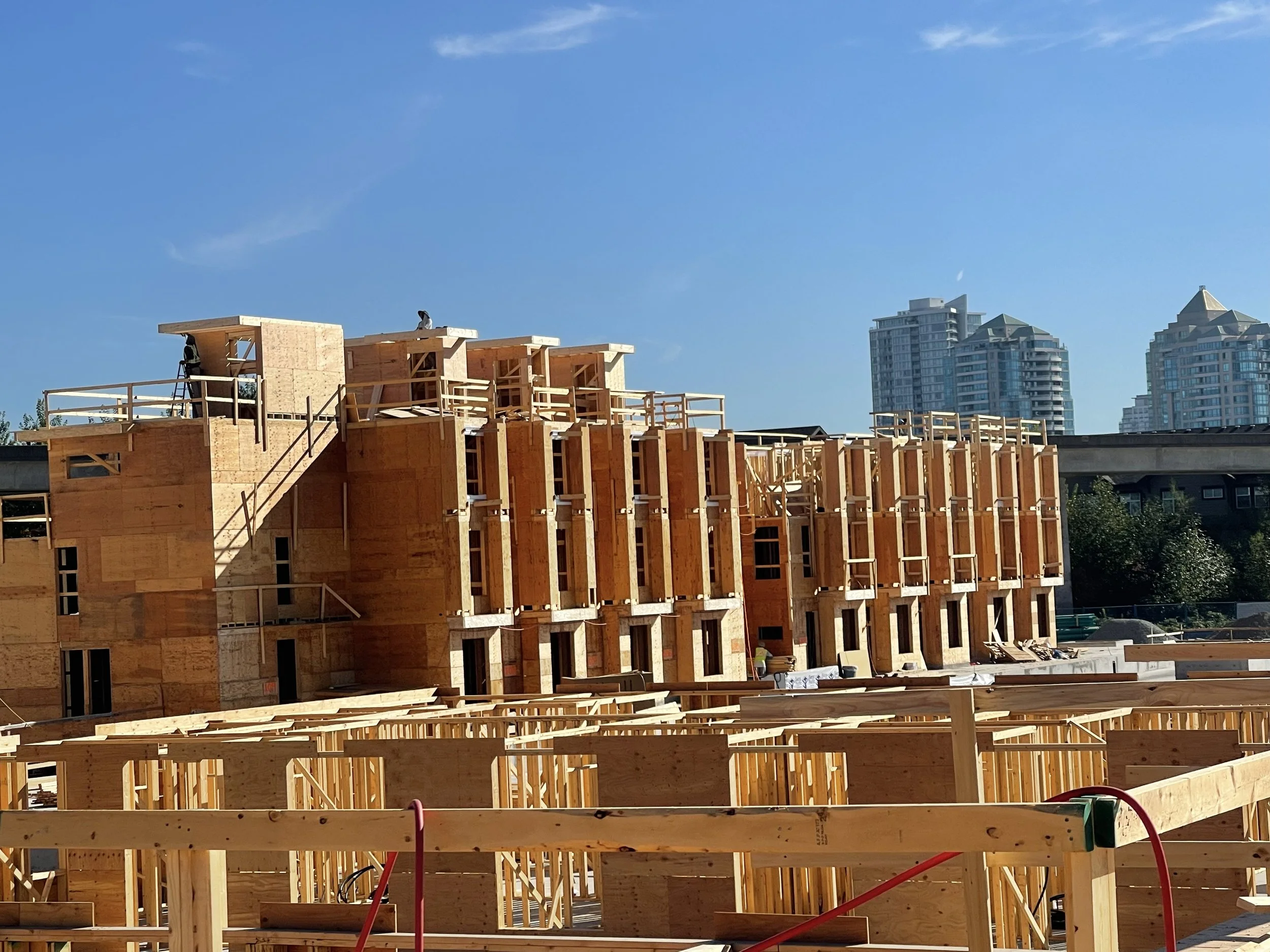 A construction site with a wooden framework of a building in progress, with tall modern buildings in the background and a clear blue sky.