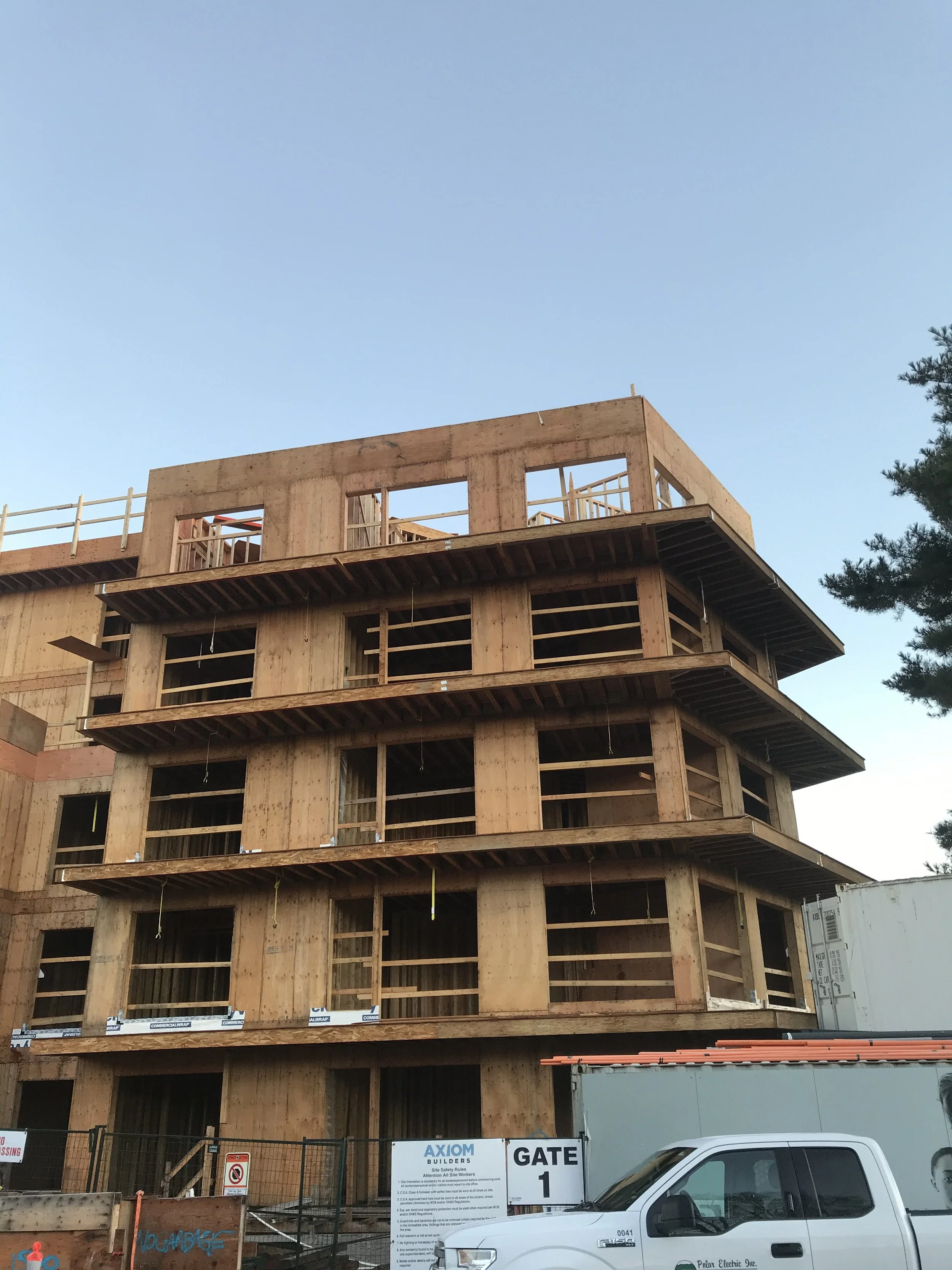 A multi-story building under construction with wooden framing, surrounded by construction signs, fencing, and a white utility truck parked in front.