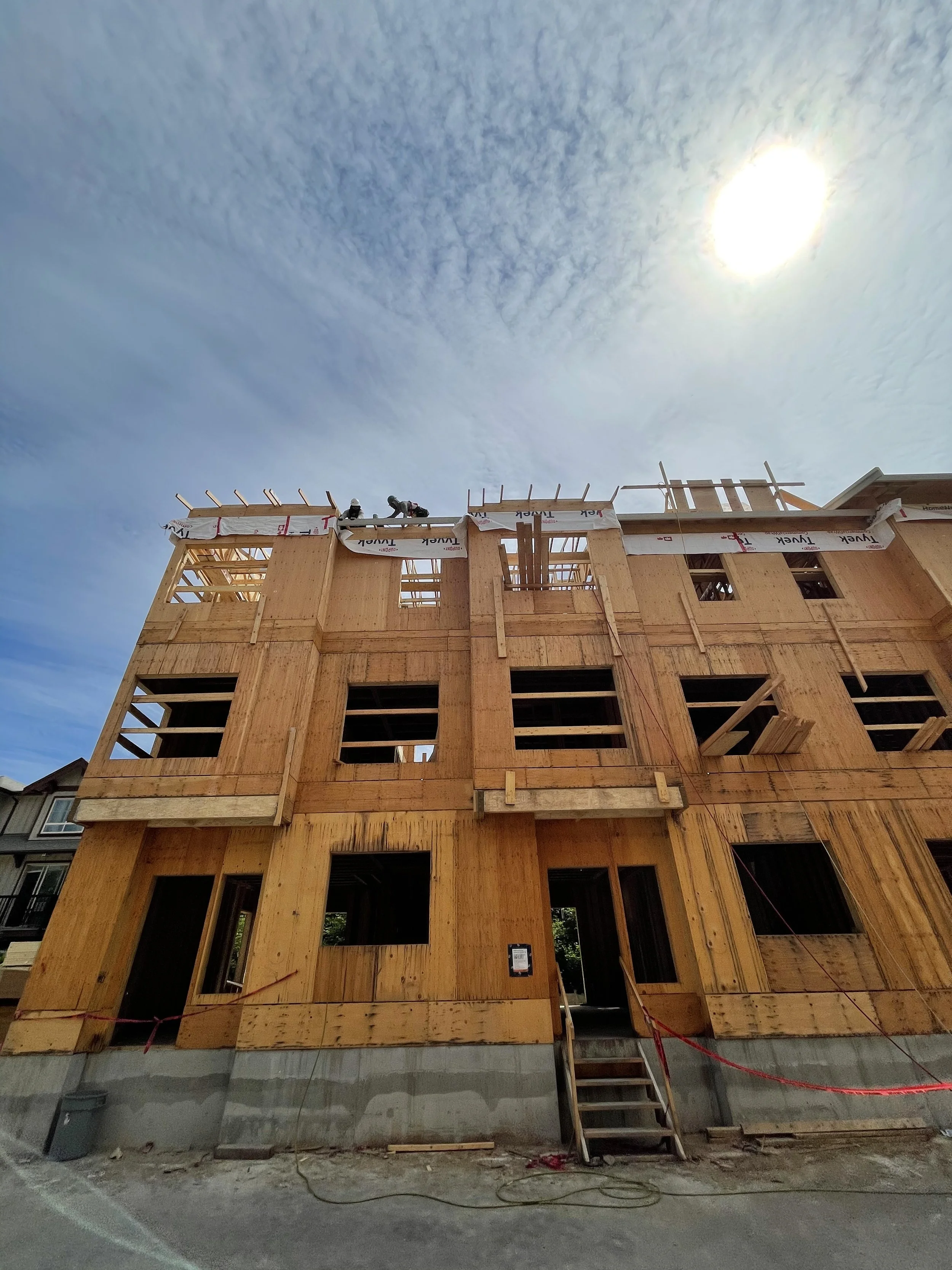 Wooden frame of a building under construction with two workers on the roof, the sky is blue with scattered clouds and the sun shining.