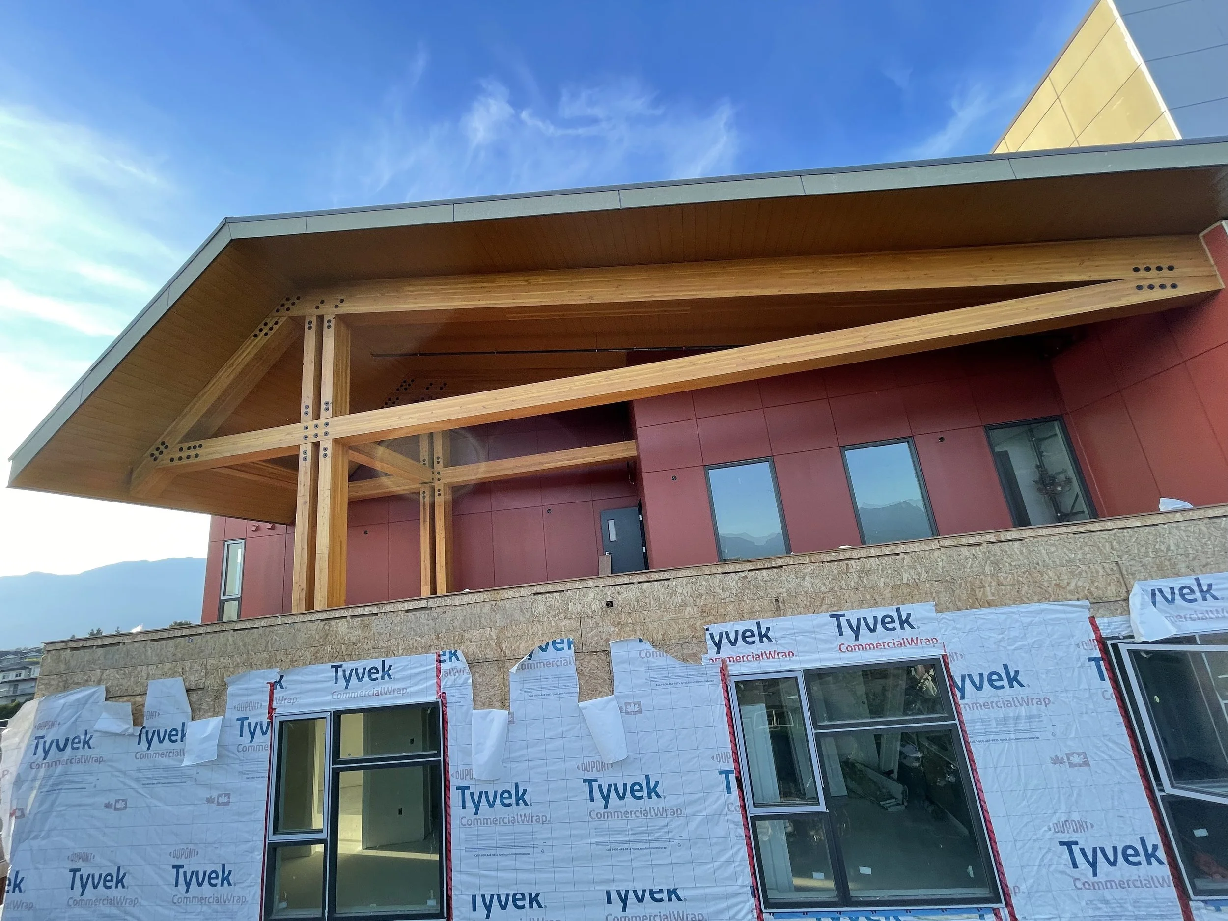 A building under construction with exterior walls partially covered in white Tyvek weather barrier and large windows installed. The upper part of the building features wood framing and a sloped roof. The construction site is outdoors with blue sky and mountains in the background.