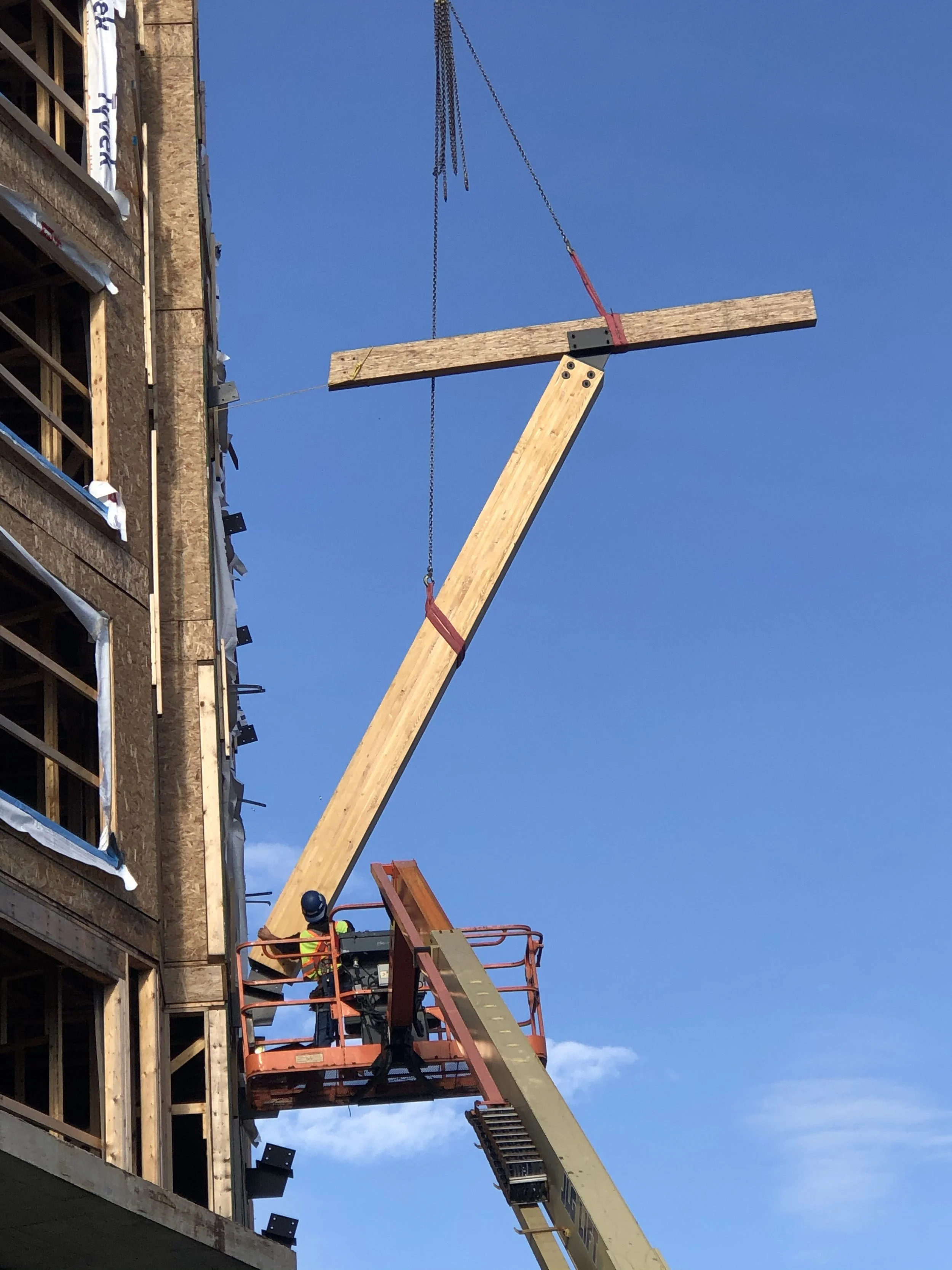 Construction worker operating a boom lift near a building under construction, with wooden beams and framing visible, against a blue sky.
