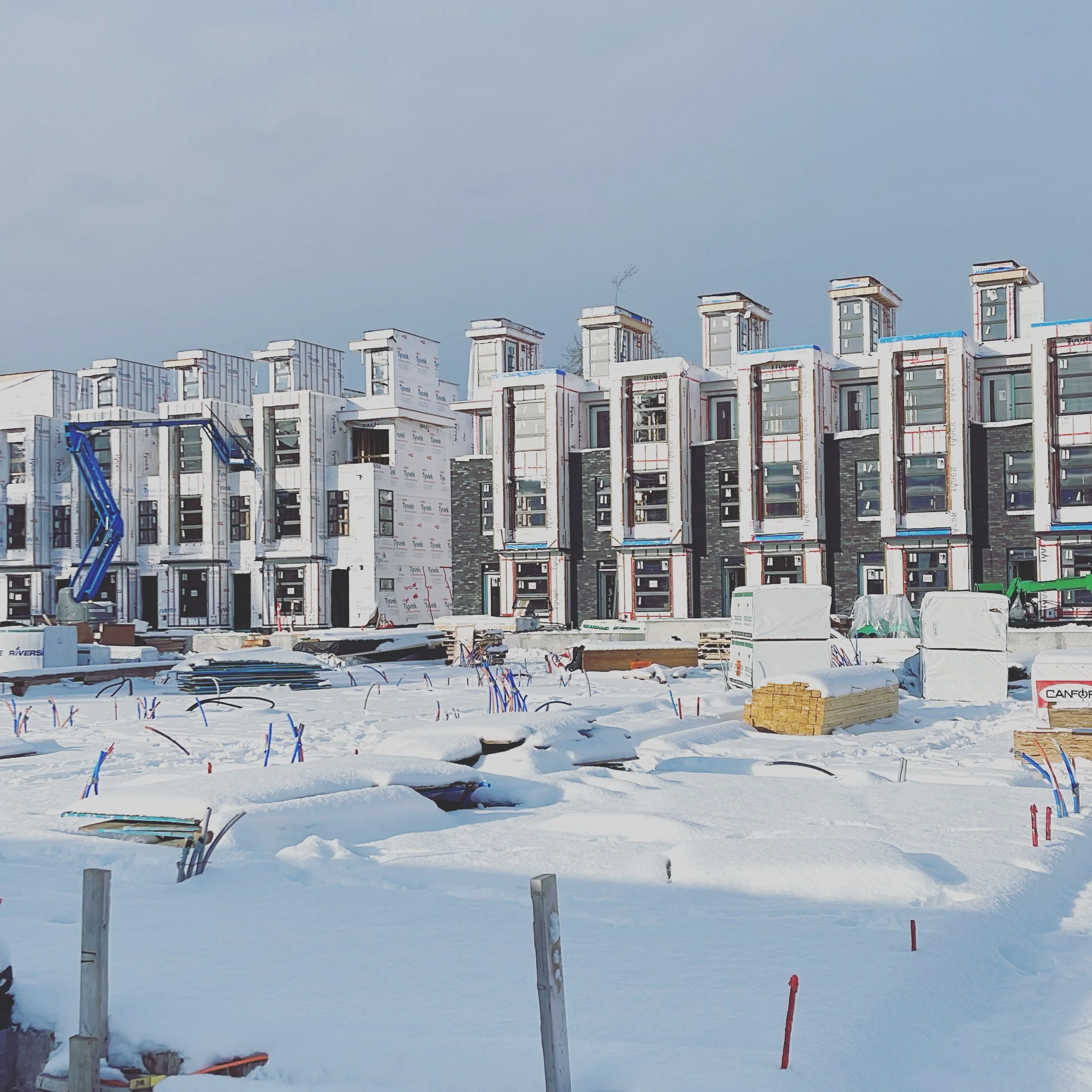 Construction site of a modern building covered in white weather-resistant sheathing, with a clear blue sky and snow on the ground.