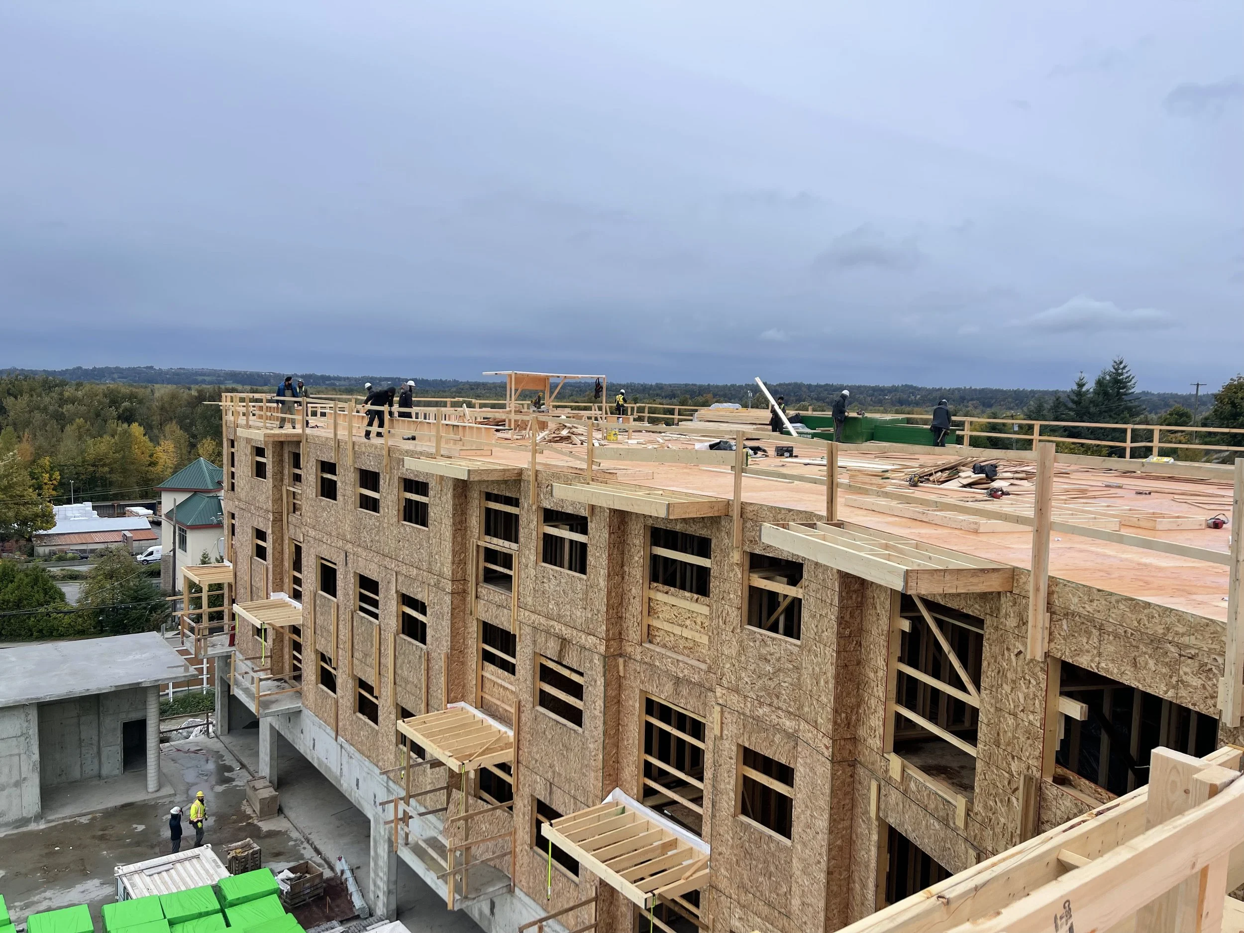 Construction workers building the roof of a multi-story wooden building with scaffolding and materials on a cloudy day.