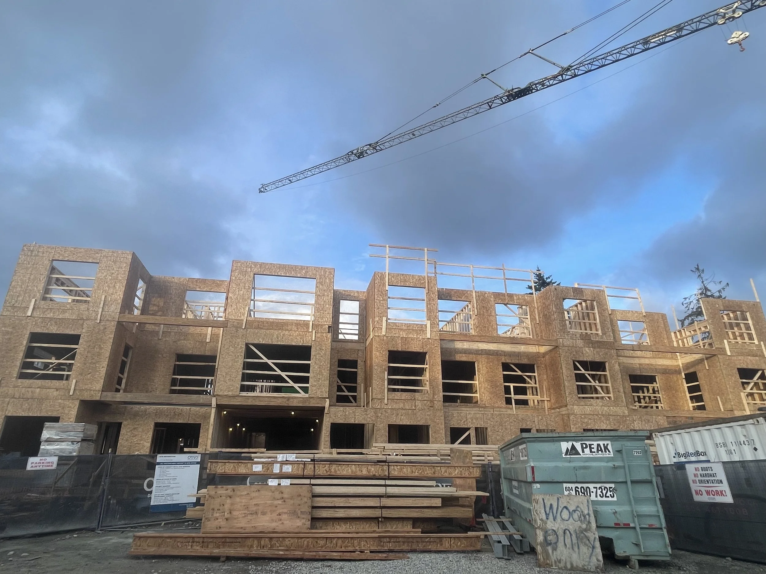 Construction site with a partially built multi-story building framework and a crane overhead. Construction materials and equipment are visible in the foreground.