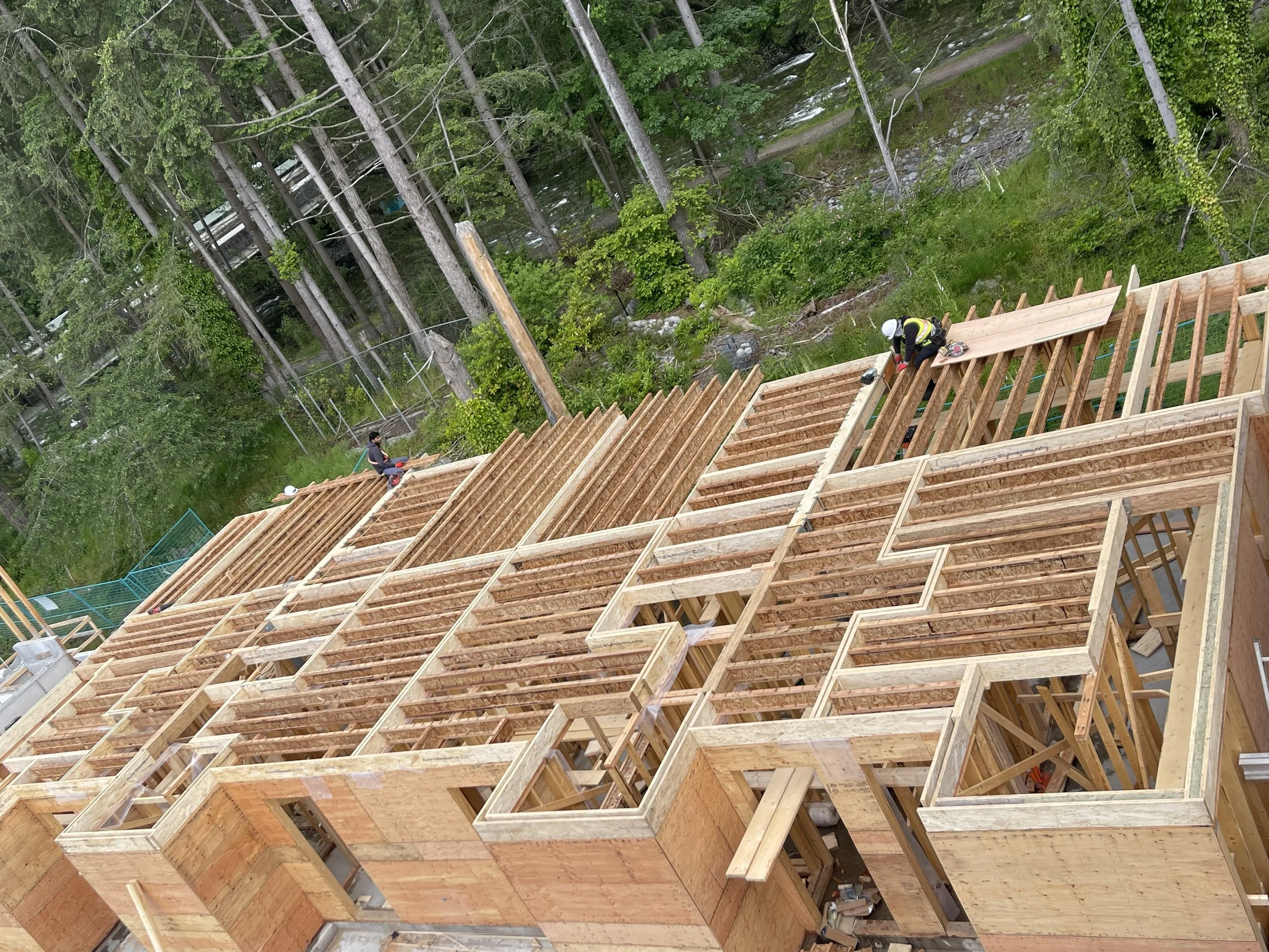 Construction workers building a multi-story wooden house frame in a wooded area next to a river or creek, with green trees and utility poles visible in the background.