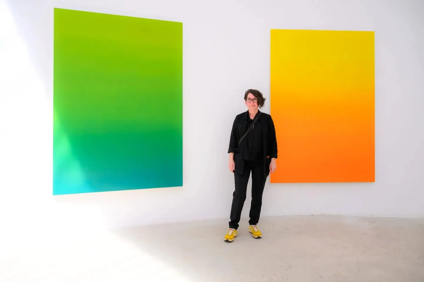 Artist Yaima Carrazana standing between two large green and orange gradient paintings.
