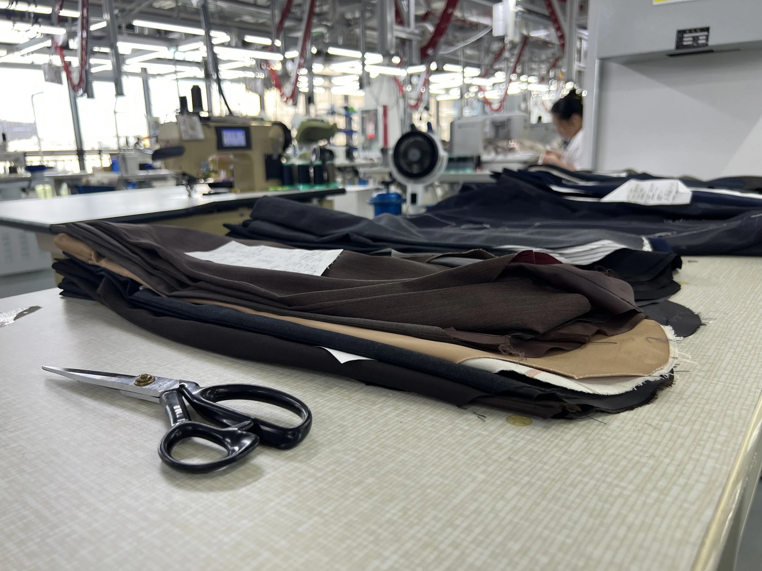 Table with neatly stacked fabric swatches, a pair of scissors, and various sewing tools in an industrial sewing or tailoring workshop.