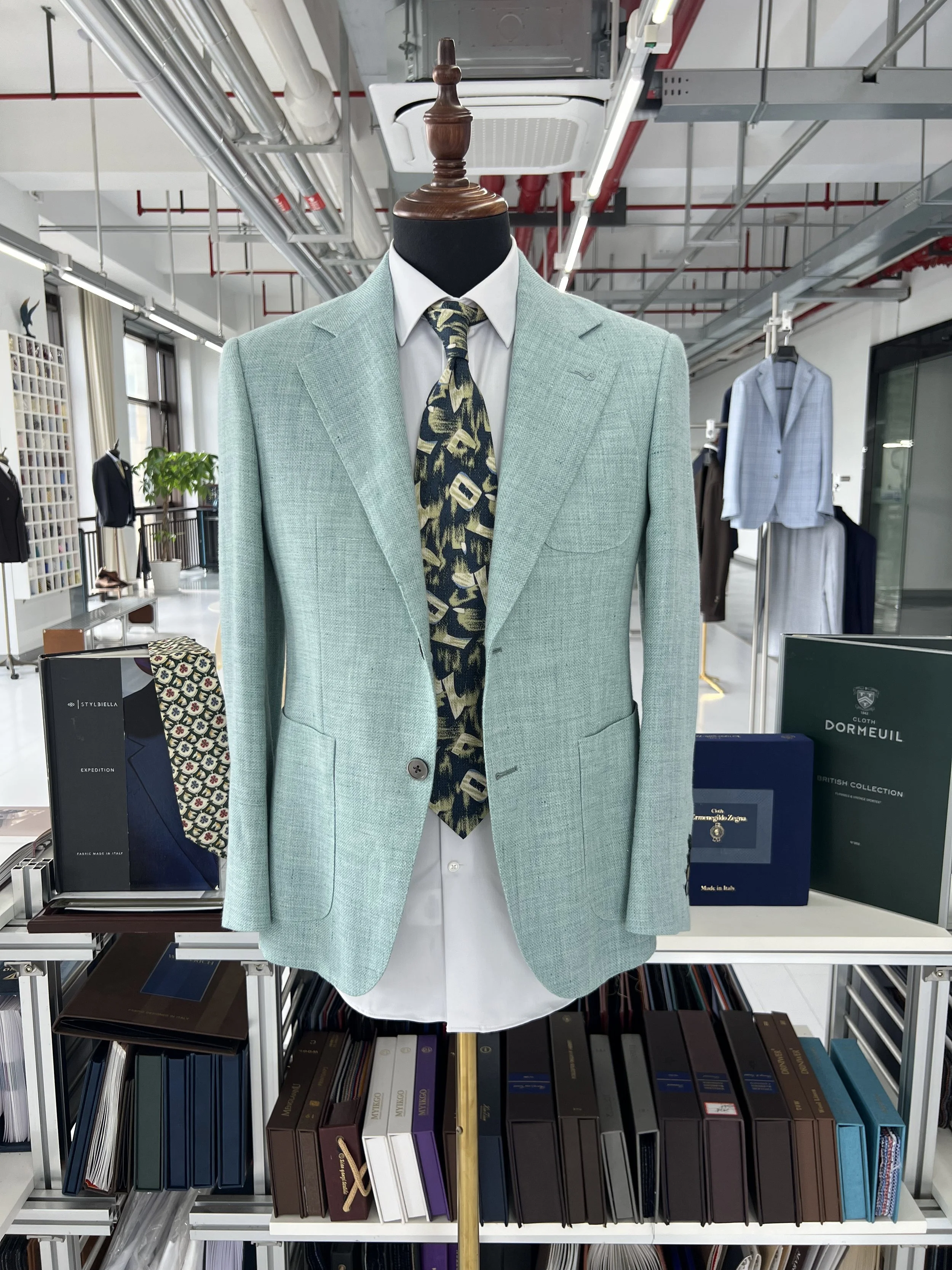 A mannequin dressed in a light green blazer, white shirt, and a patterned tie, displayed in a store with shelves of books and clothing in the background.
