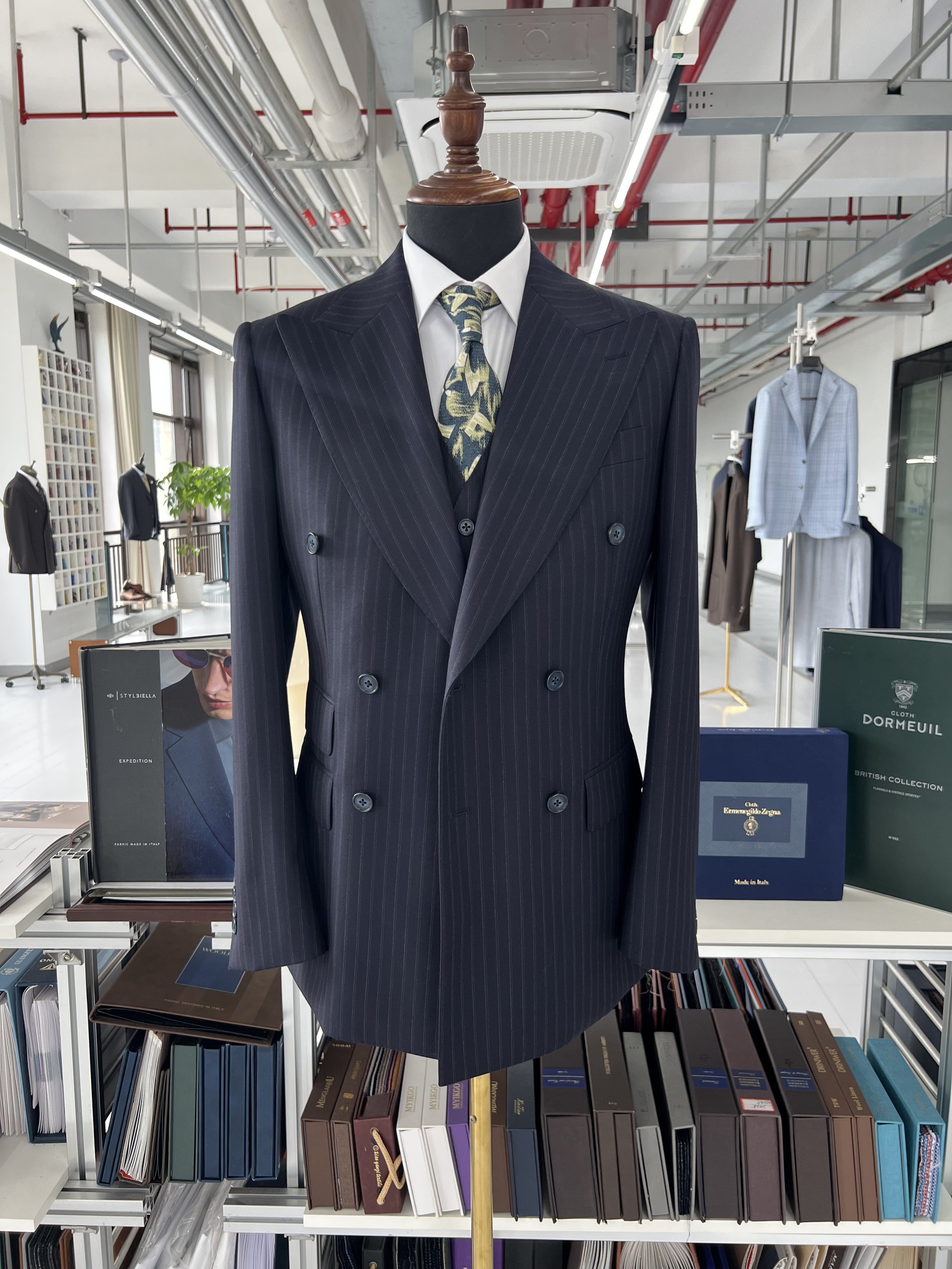 Mannequin wearing a navy blue pinstripe blazer, white shirt, and floral tie, displayed in a clothing store. The store has books and clothing on shelves, and large windows in the background.