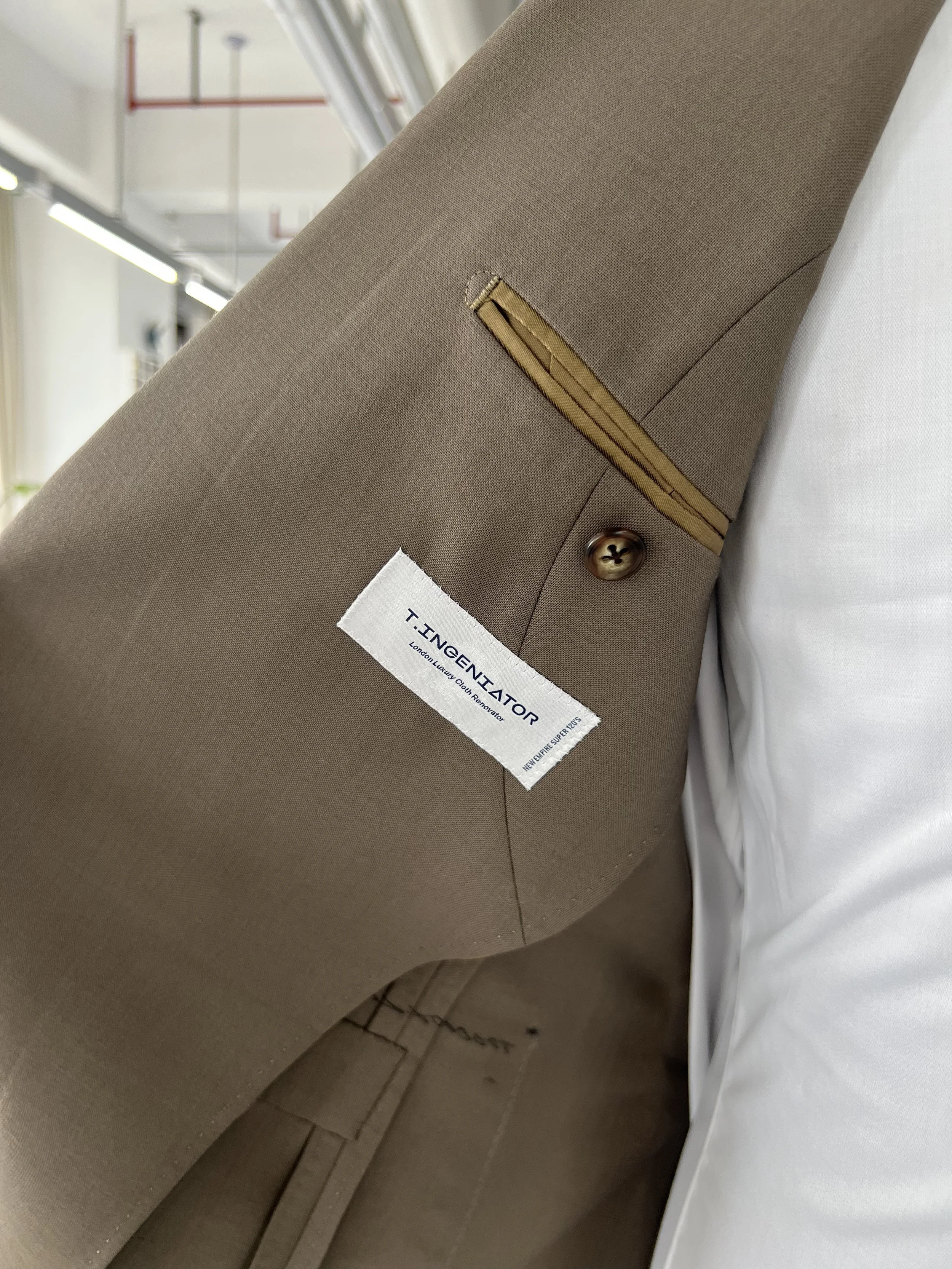 Close-up of a beige suit jacket sleeve with a gold zipper and a brown button, with a white label that reads 'T. INGENTATOR, London Luxury Cloth Renovator' attached to the sleeve. The background shows a white surface and part of a tiled floor.