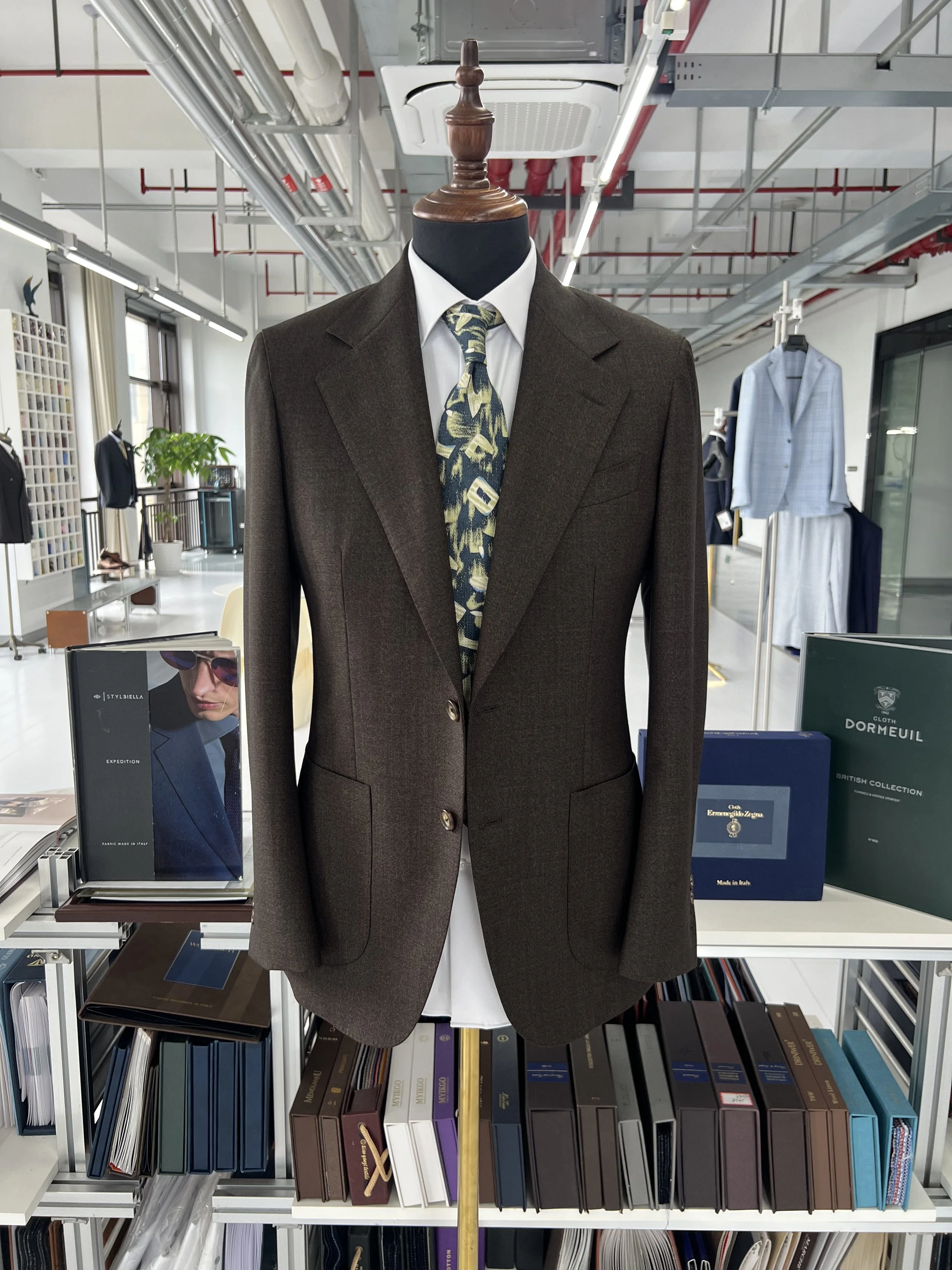 Mannequin dressed in a brown suit jacket with a white shirt and a patterned tie, on a wooden stand in an office or showroom with bookshelves and clothing in the background.