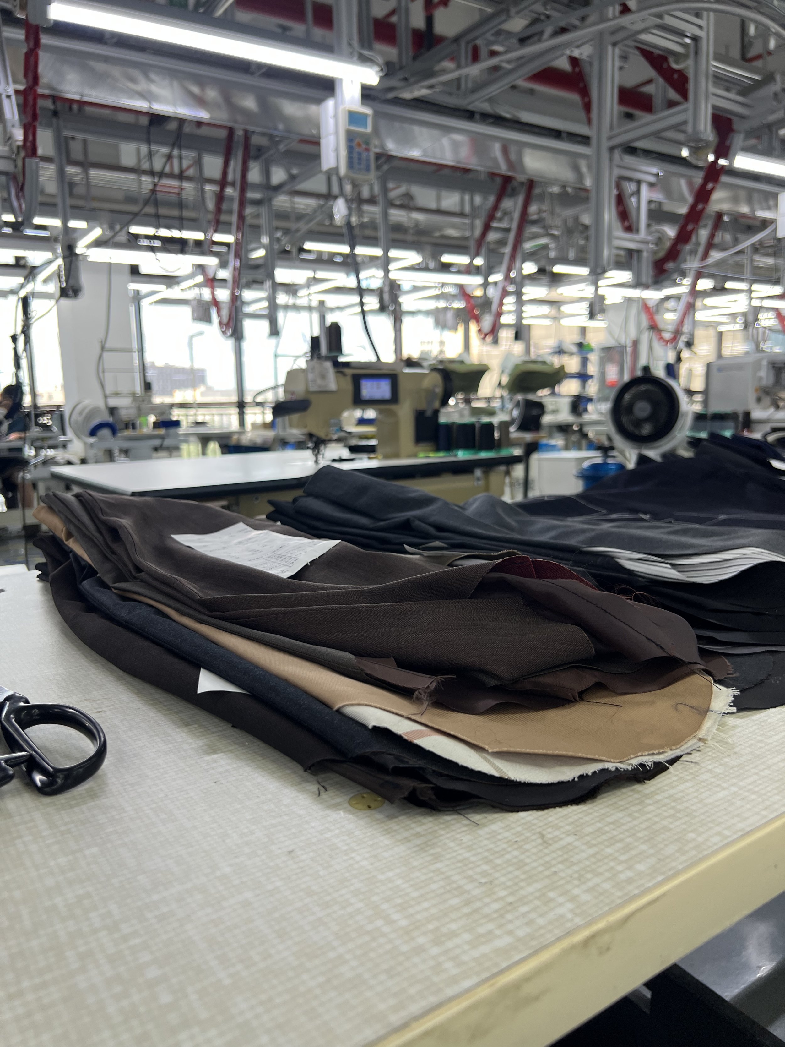 Layers of fabric and paper on a light-colored table at a sewing workshop, with scissors and other sewing tools visible. In the background, there is industrial sewing equipment and workspace with metallic shelving and bright lighting.