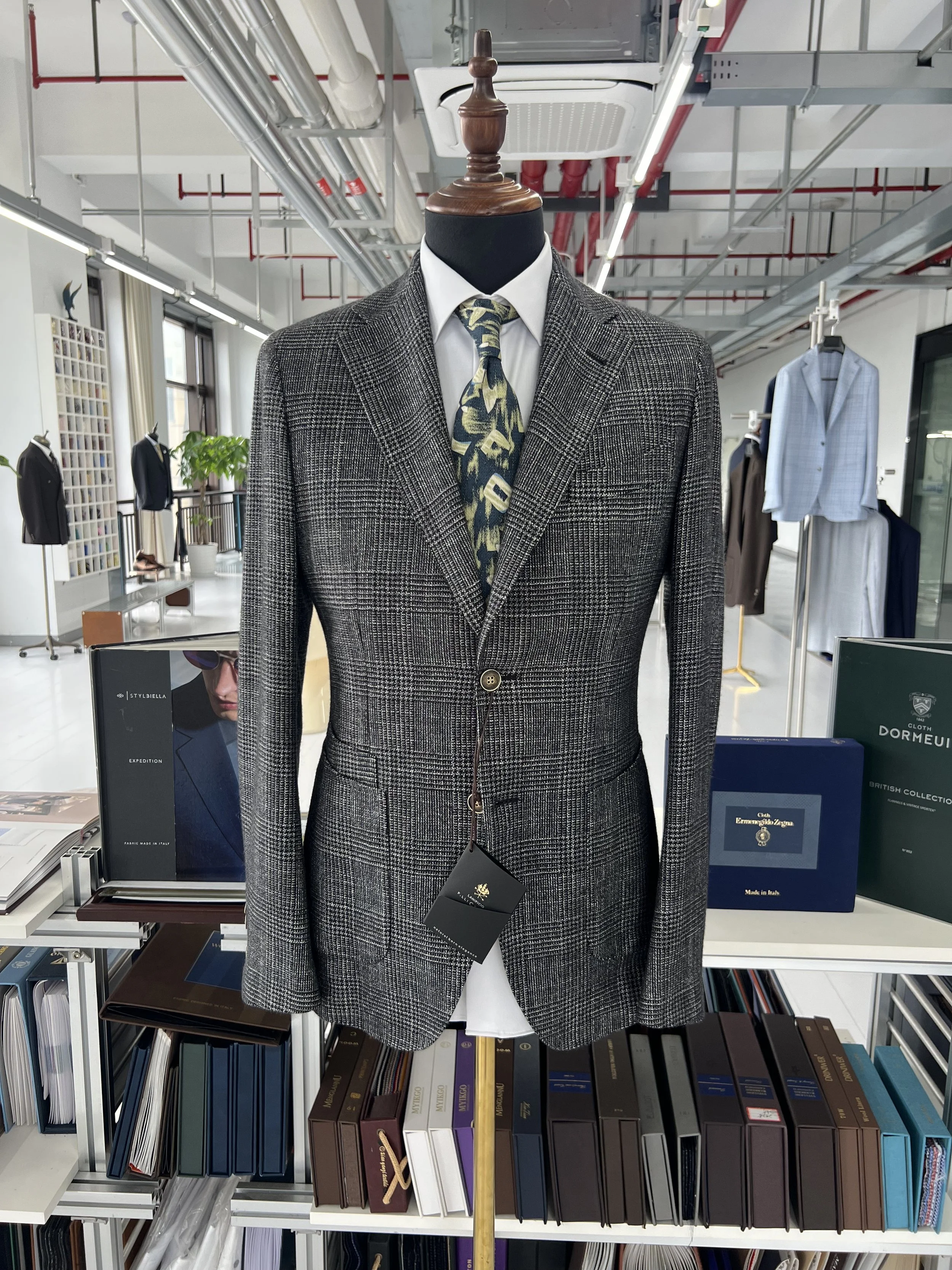 Mannequin dressed in a plaid suit jacket, white shirt, and a multicolored tie, displayed in a store with shelves of books and clothing in the background.