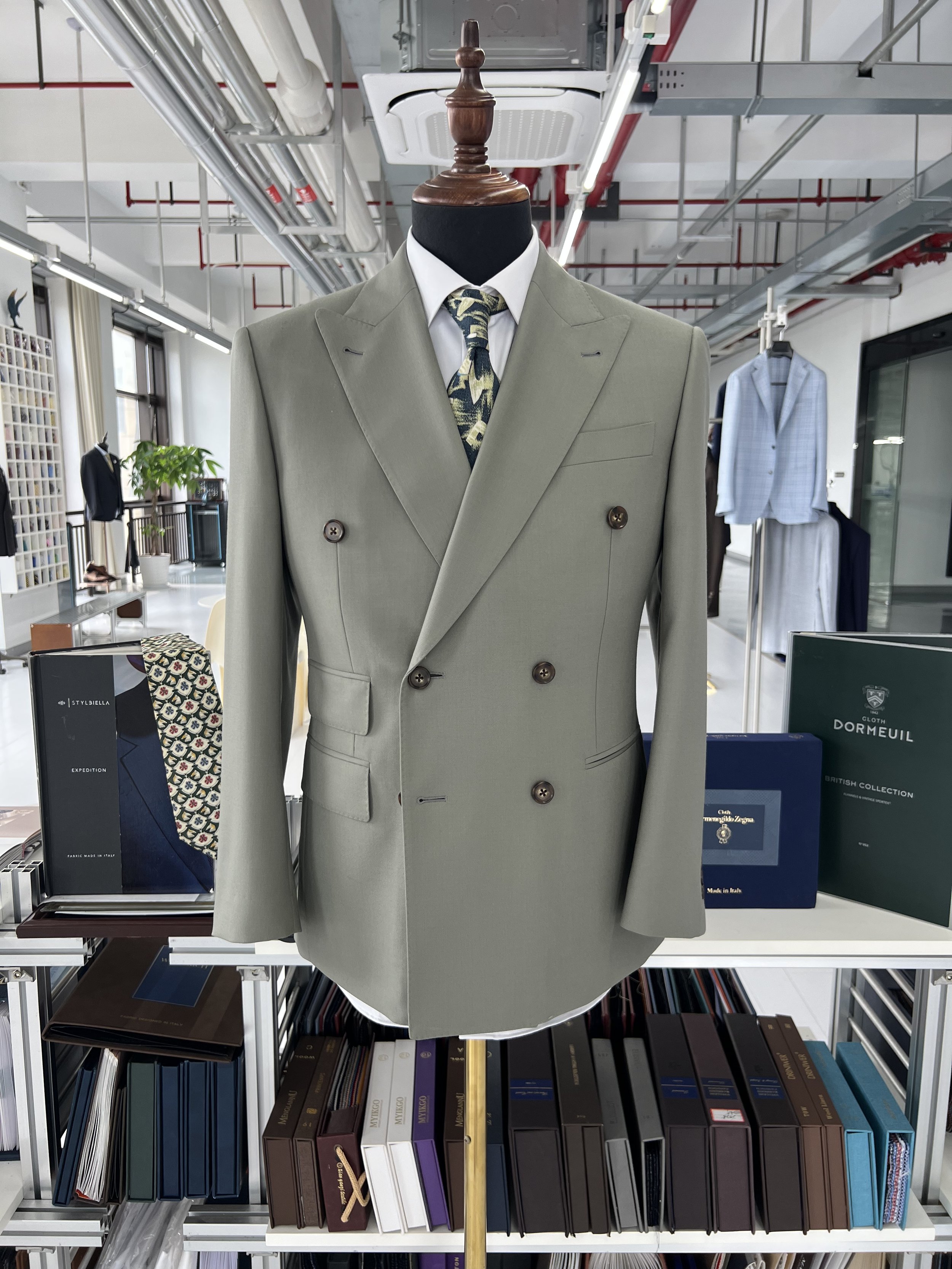 A mannequin dressed in a light gray double-breasted suit with a white shirt and a patterned tie, standing in a store with shelves of books and clothing in the background.