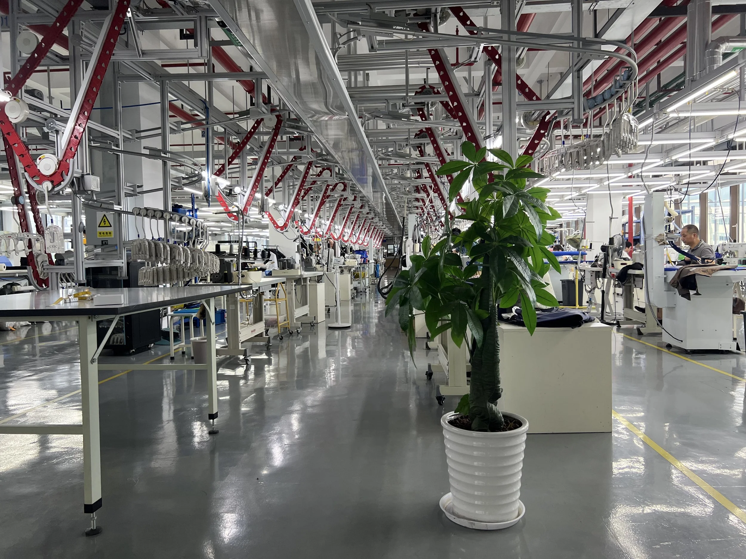 An industrial workspace with metal framework, conveyor belts, machinery, and a prominent potted green plant in the foreground.