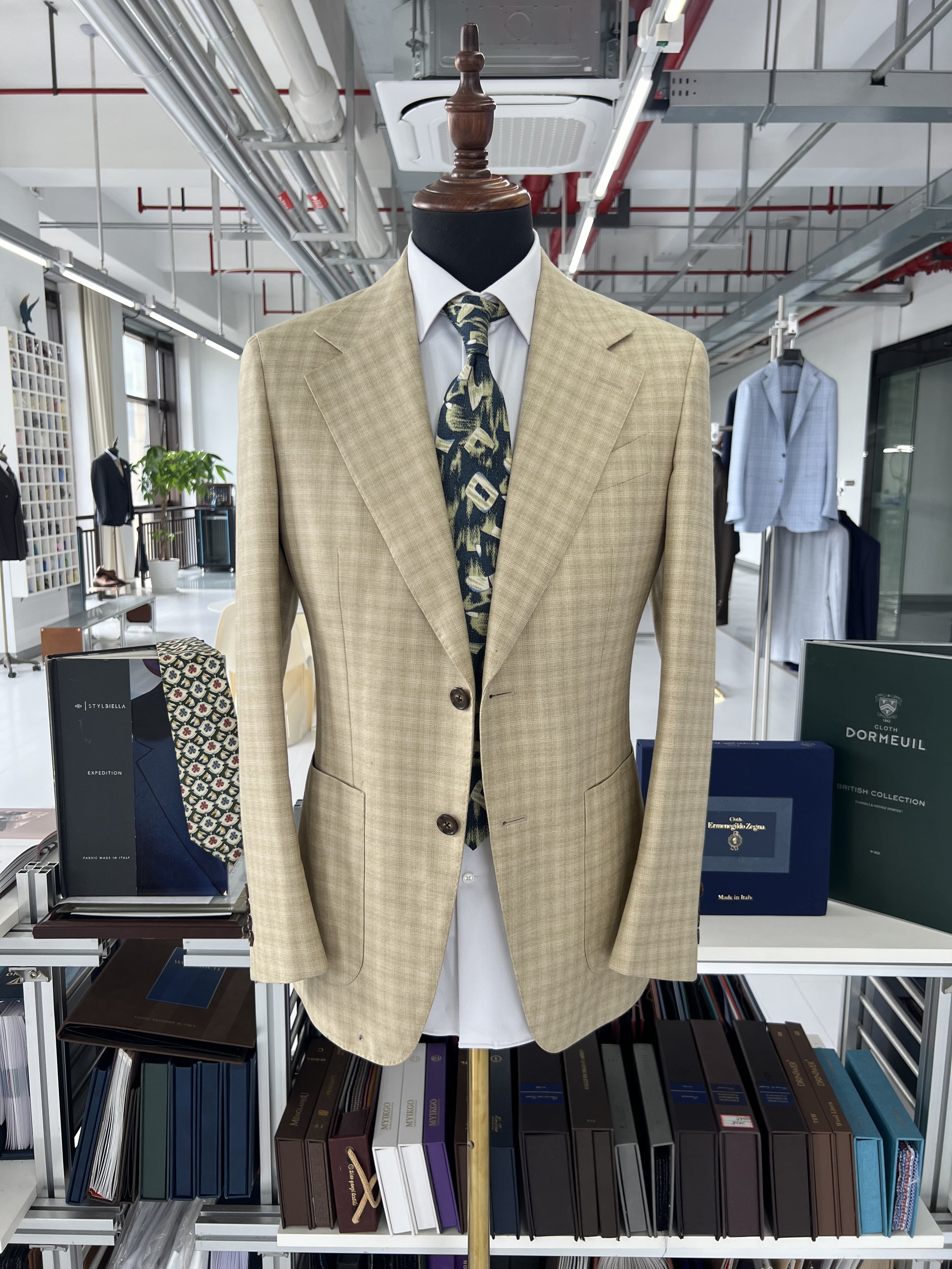 A beige suit jacket with a tie on a mannequin in a clothing store. The store has various suits and shirts on display, and shelves with books and folders in the background.