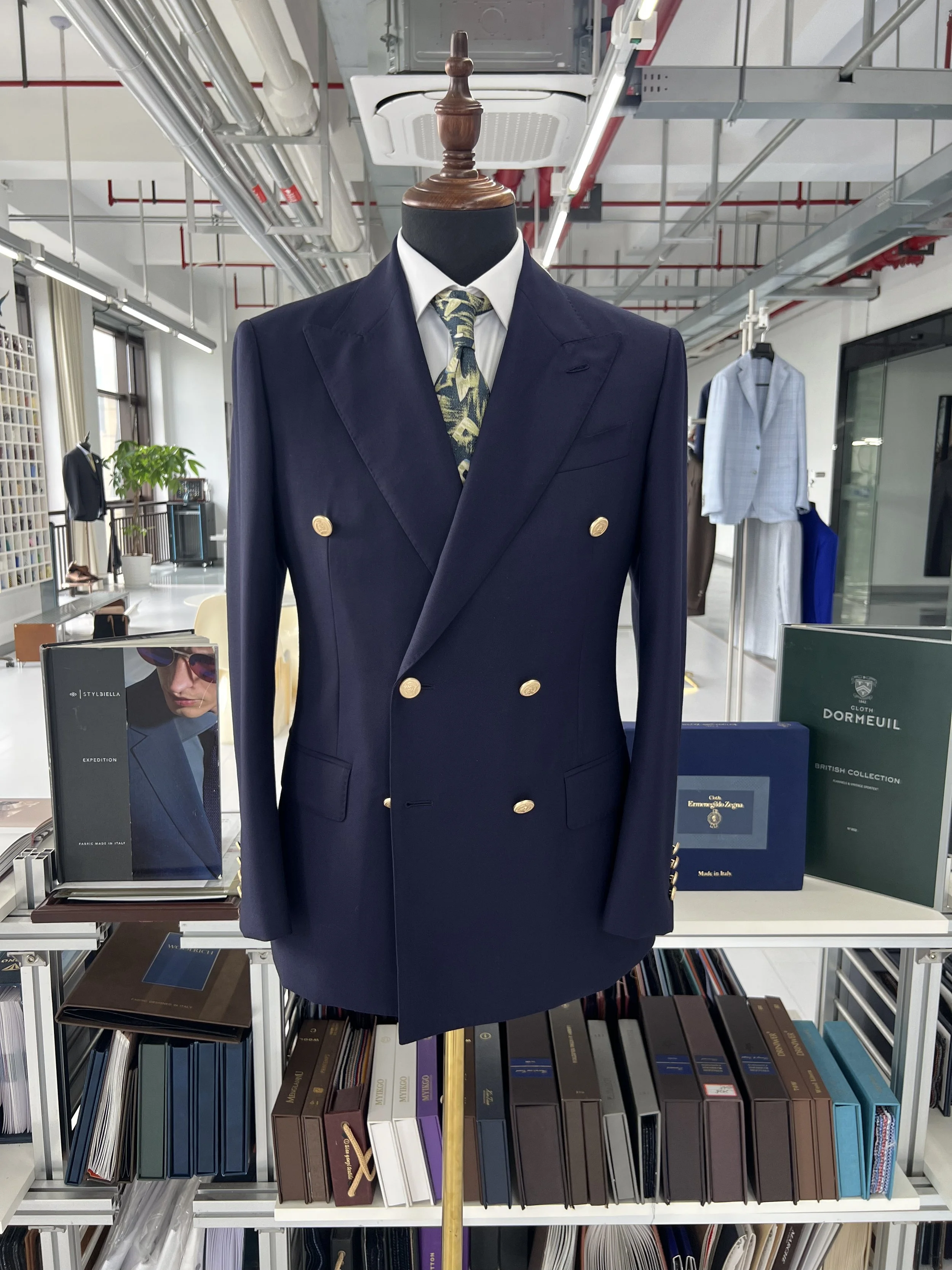 Mannequin dressed in a navy double-breasted blazer with gold buttons, white shirt, and a patterned tie, displayed on a stand in a clothing store with shelves of fabric swatches and books in the background.