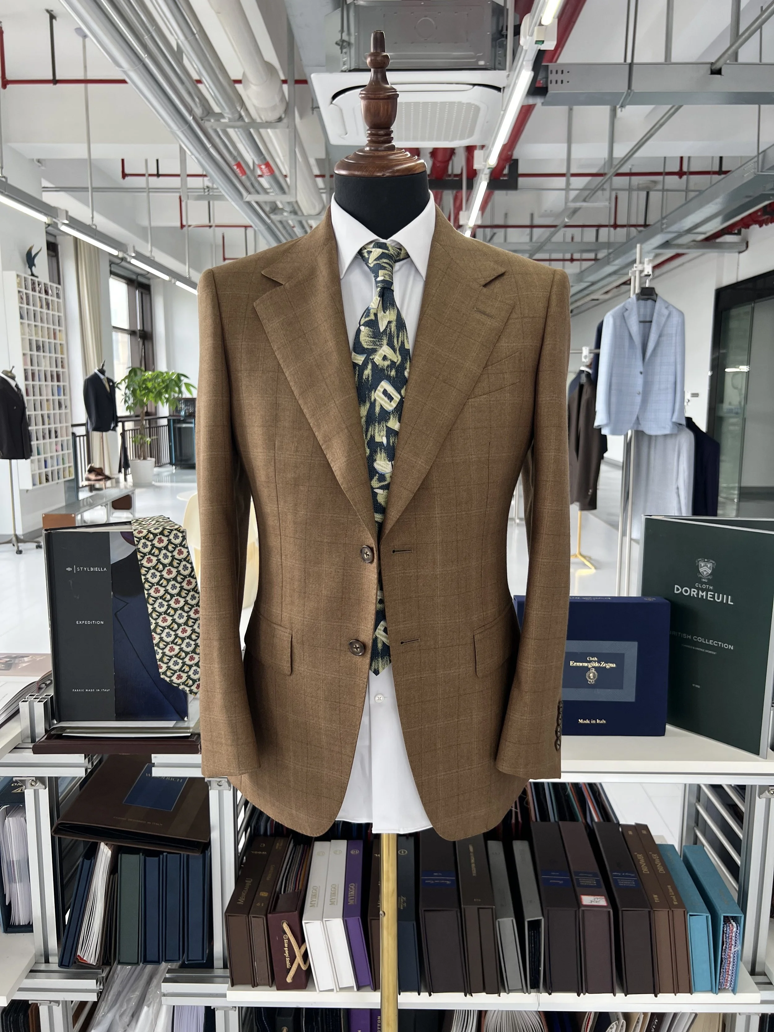 A mannequin wearing a brown suit jacket, white shirt, and patterned tie, situated in a store or showroom with shelves of fabric swatches and clothing in the background.