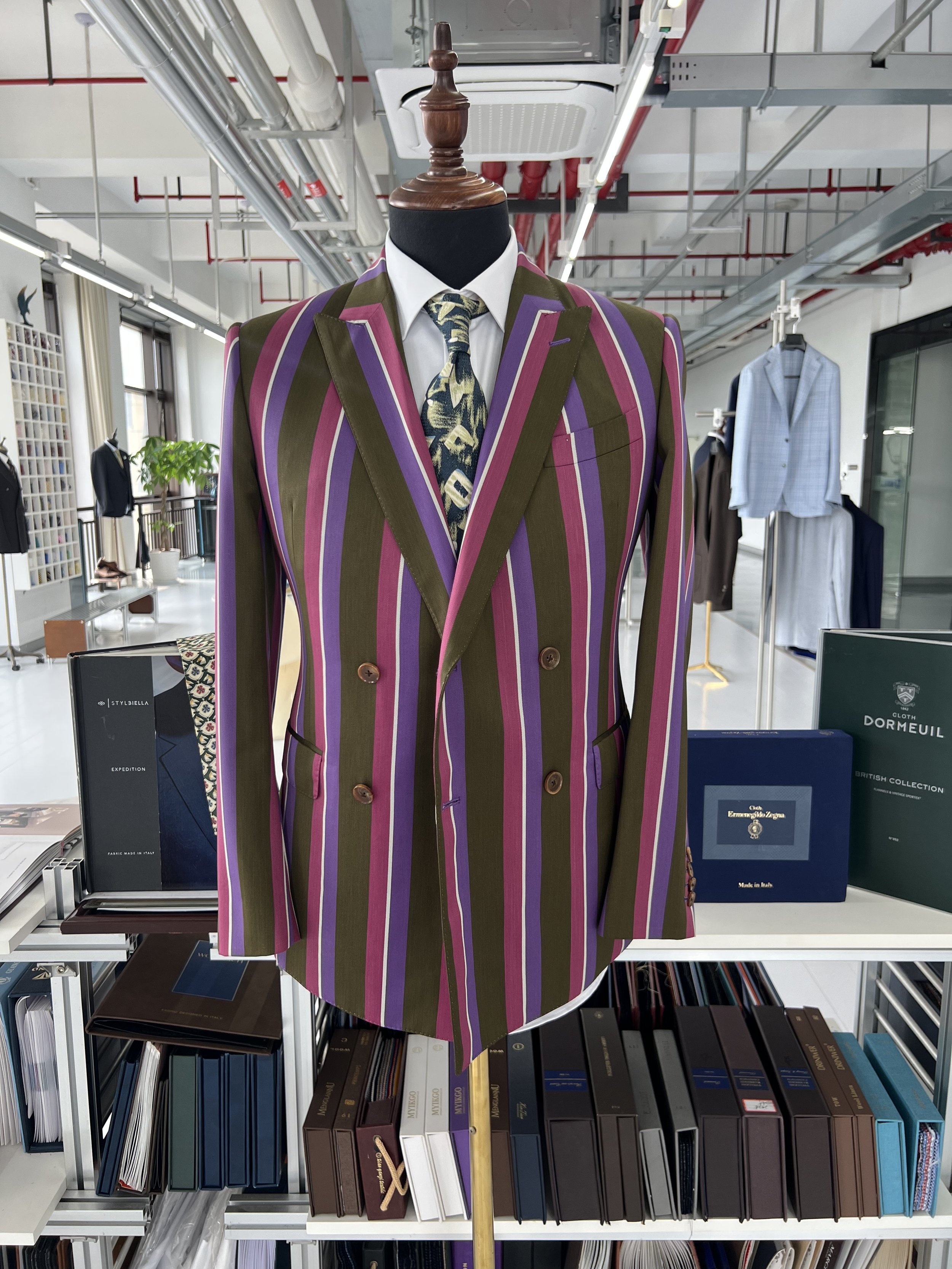 A mannequin dressed in a colorful striped blazer and a patterned tie, displayed in a store with shelves of books and clothing in the background.