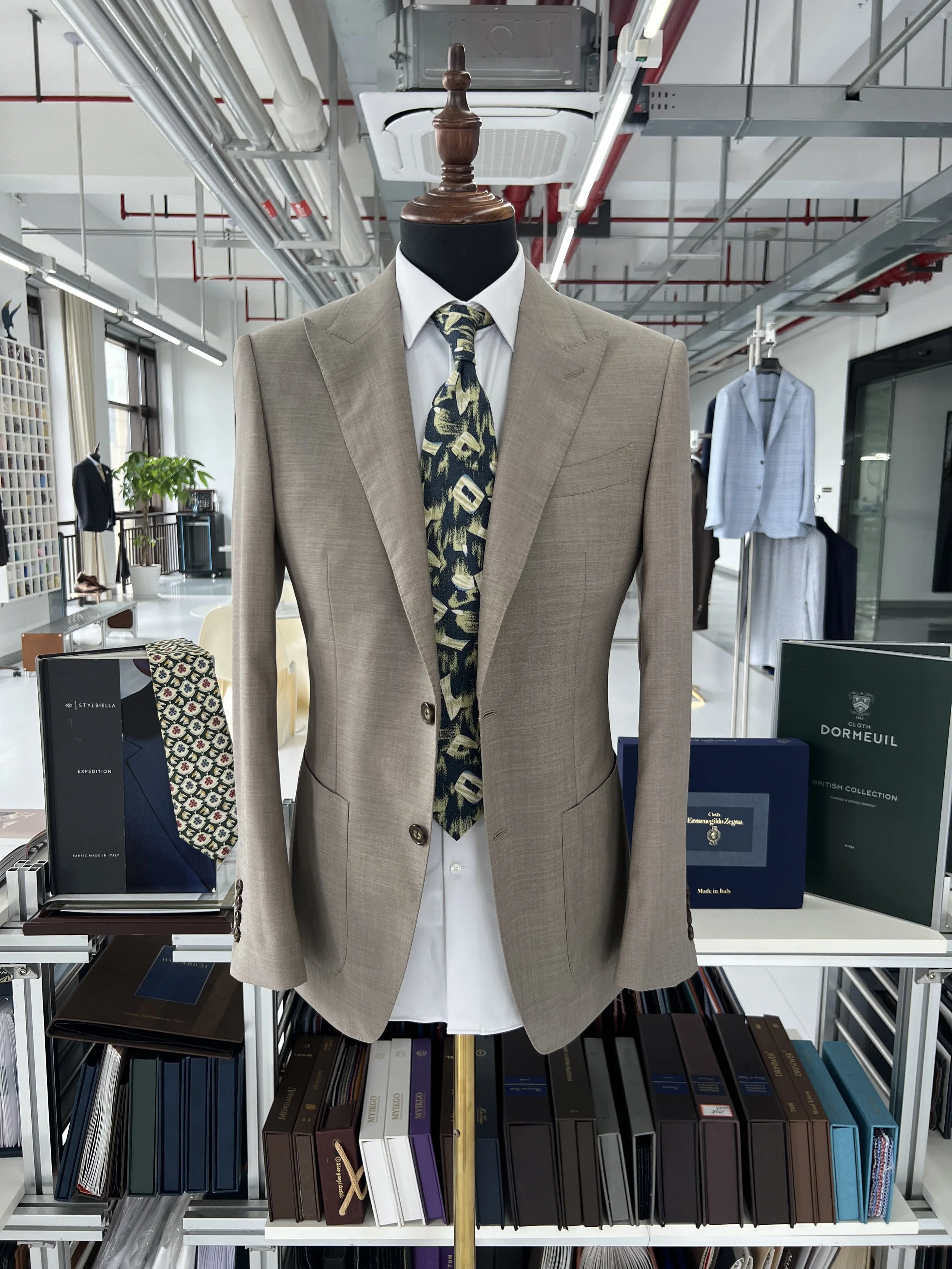 A men's beige suit displayed on a dress form mannequin with a patterned tie, in a retail store. Shelves behind hold books, boxes, and clothing items.