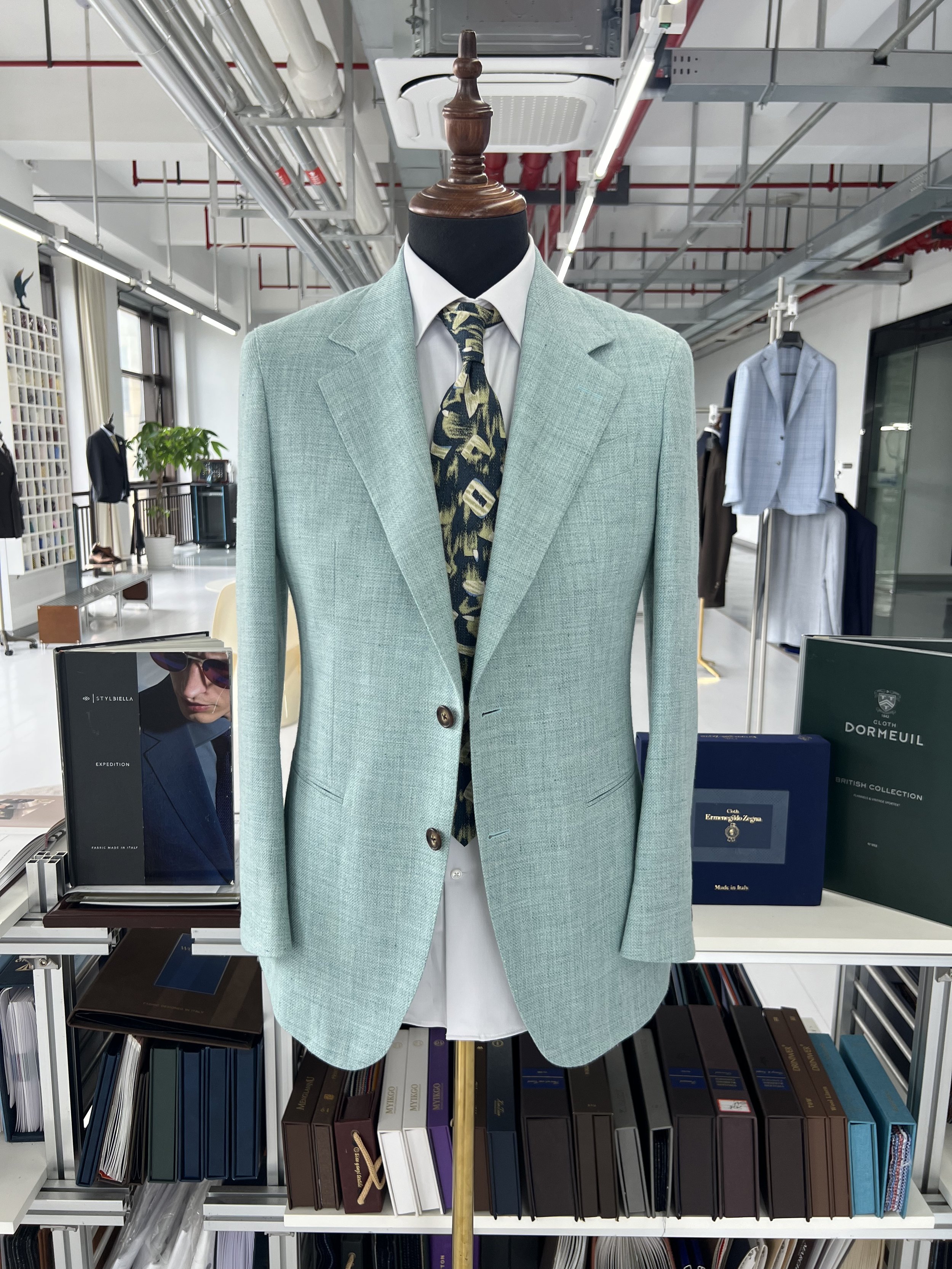 A mannequin dressed in a light green blazer, white shirt, and a patterned tie, displayed in a clothing store with shelves of books and other clothing items visible in the background.