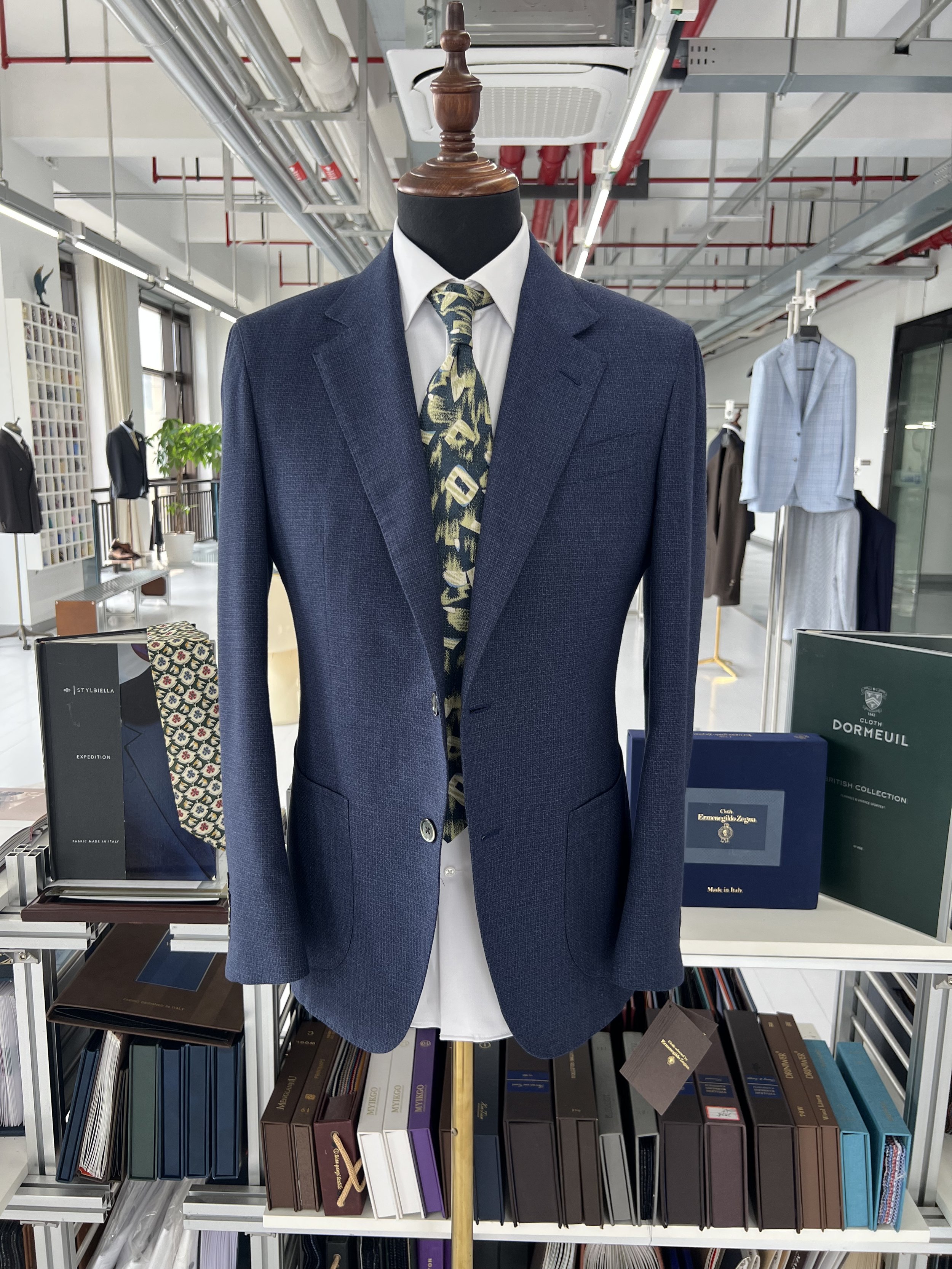 Mannequin dressed in a dark blue suit jacket, white shirt, and a patterned tie, displayed in a clothing store with shelves of books and additional garments in the background.