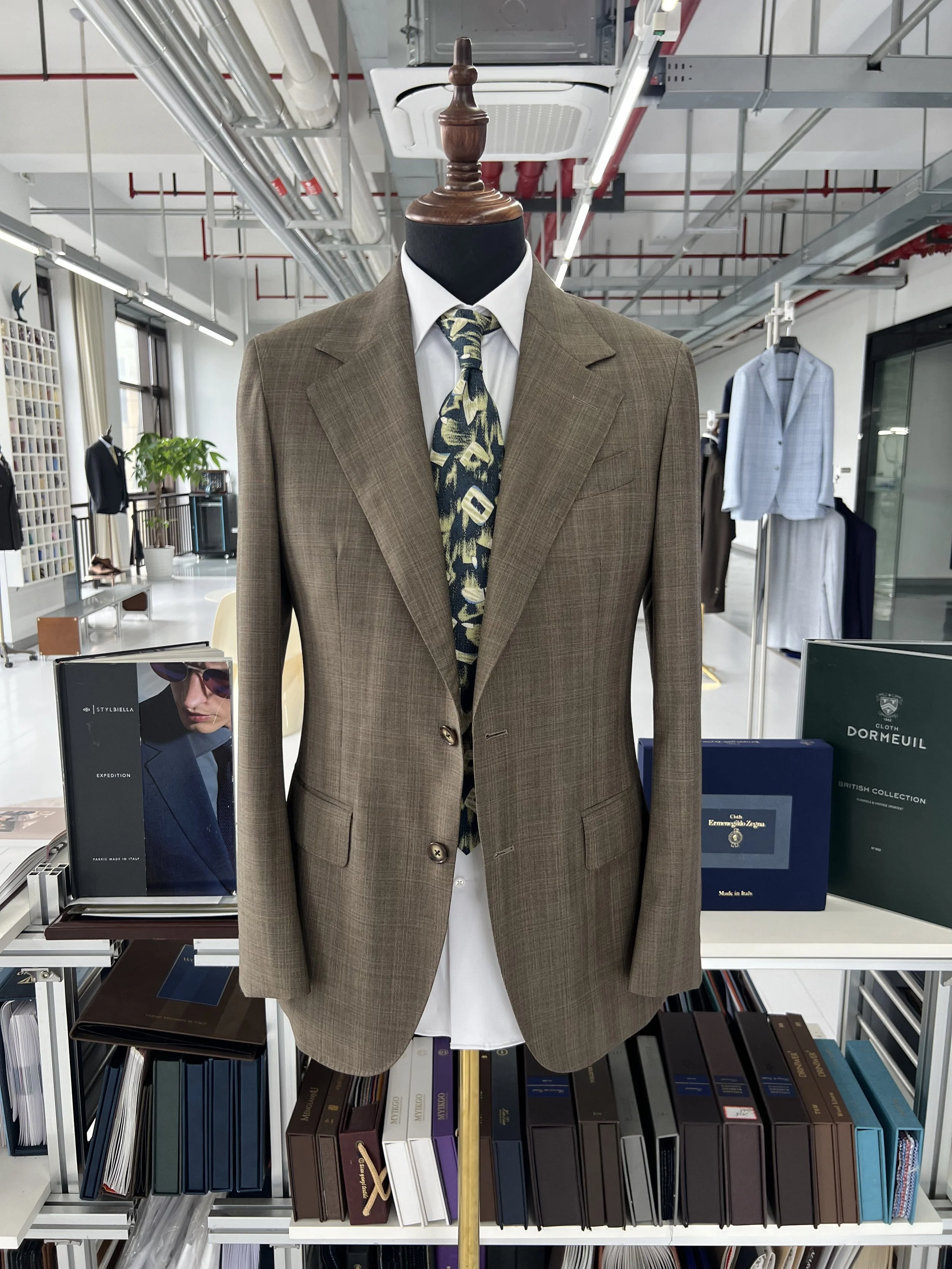 Mannequin dressed in a brown plaid suit with a white shirt and a patterned tie, on display in a store or showroom with shelves of books and fashion items in the background.