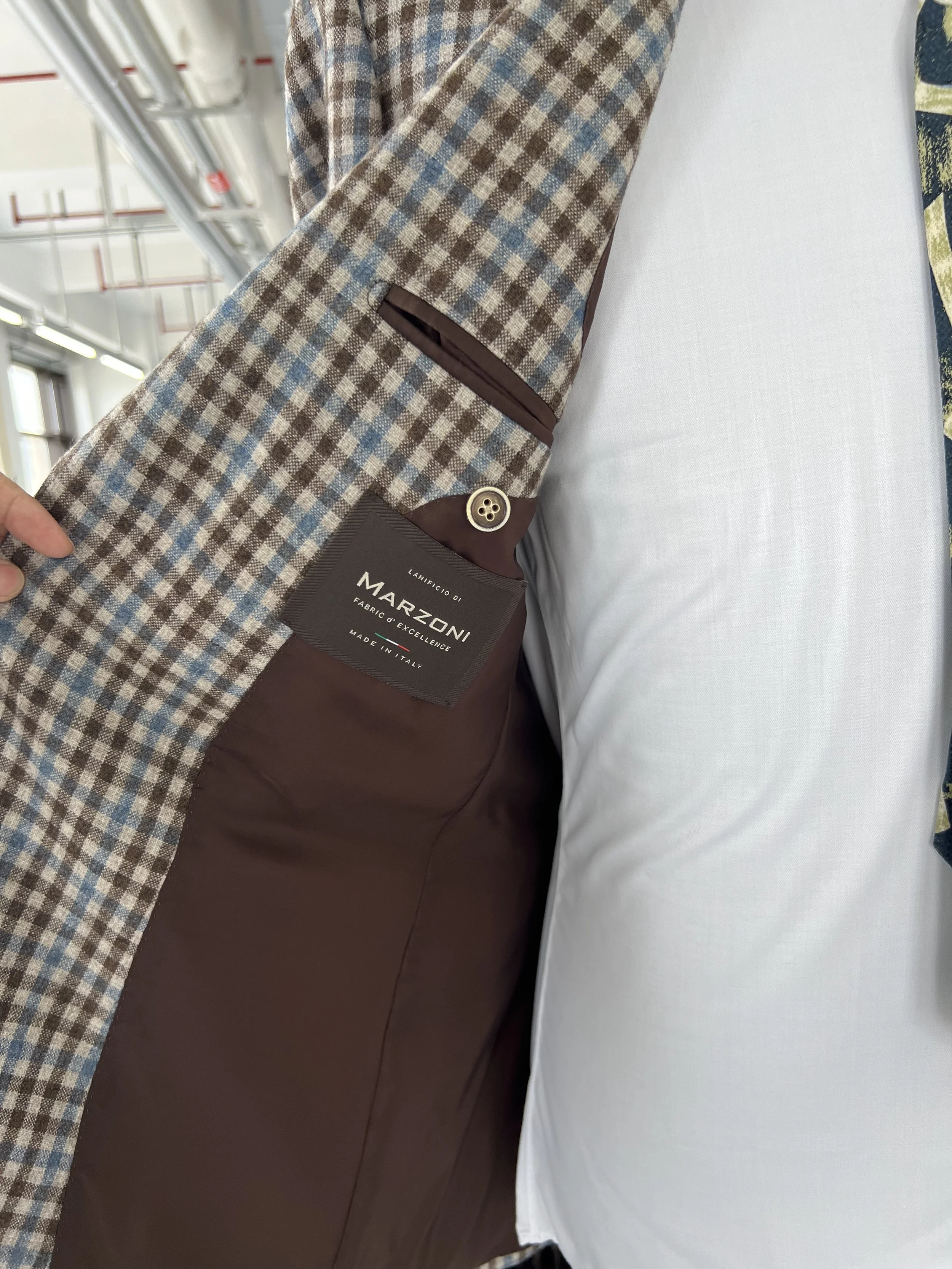 Close-up of a men's blazer with a checkered pattern and a tag that reads 'Lanificio di Marzoni, fabric & excellence, made in Italy.' The person is wearing a white shirt.