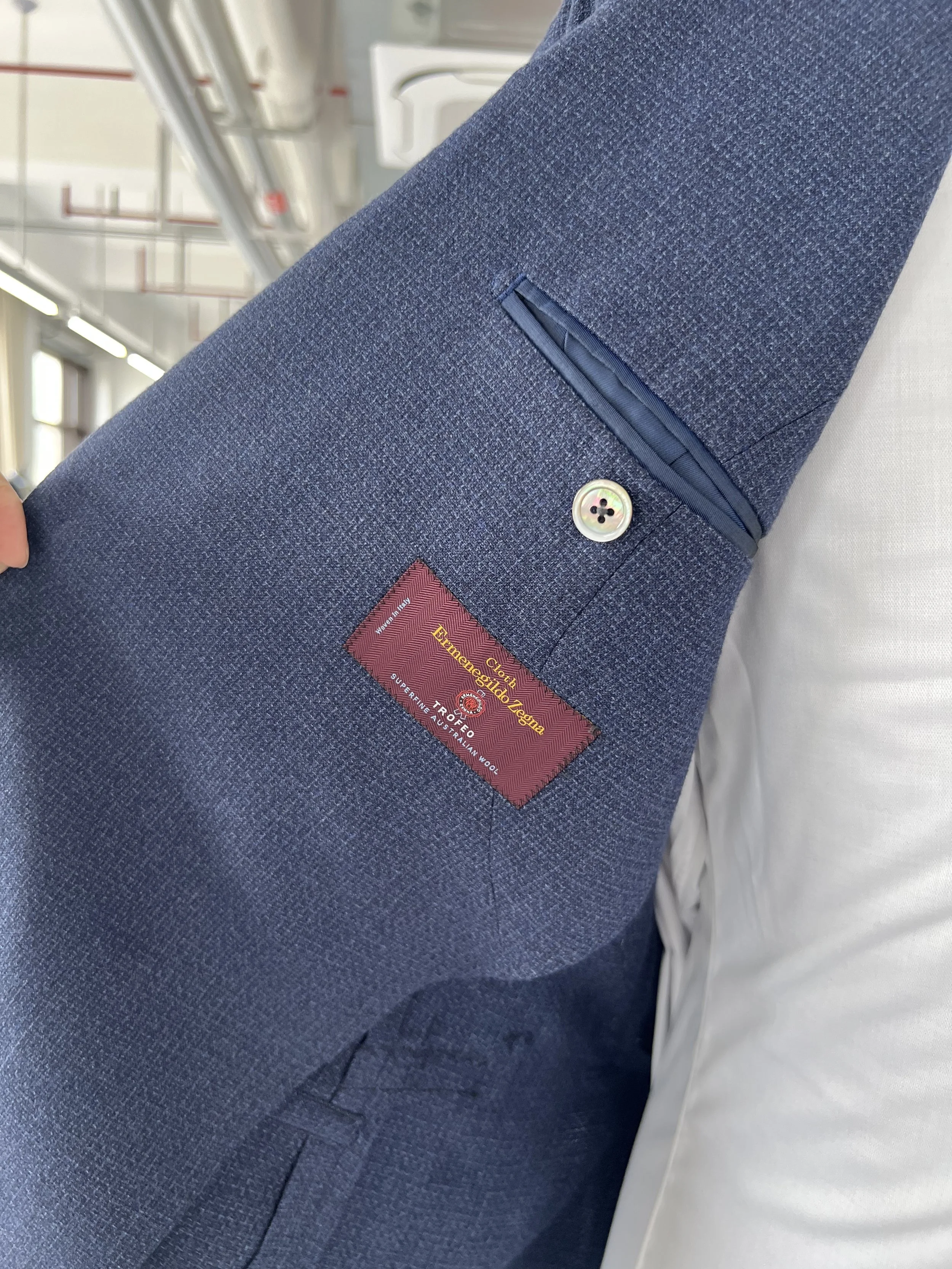 Close-up of a men's blue suit jacket's pocket on a person's arm, with a white button and a red tag reading "Superfine Australian Wool."