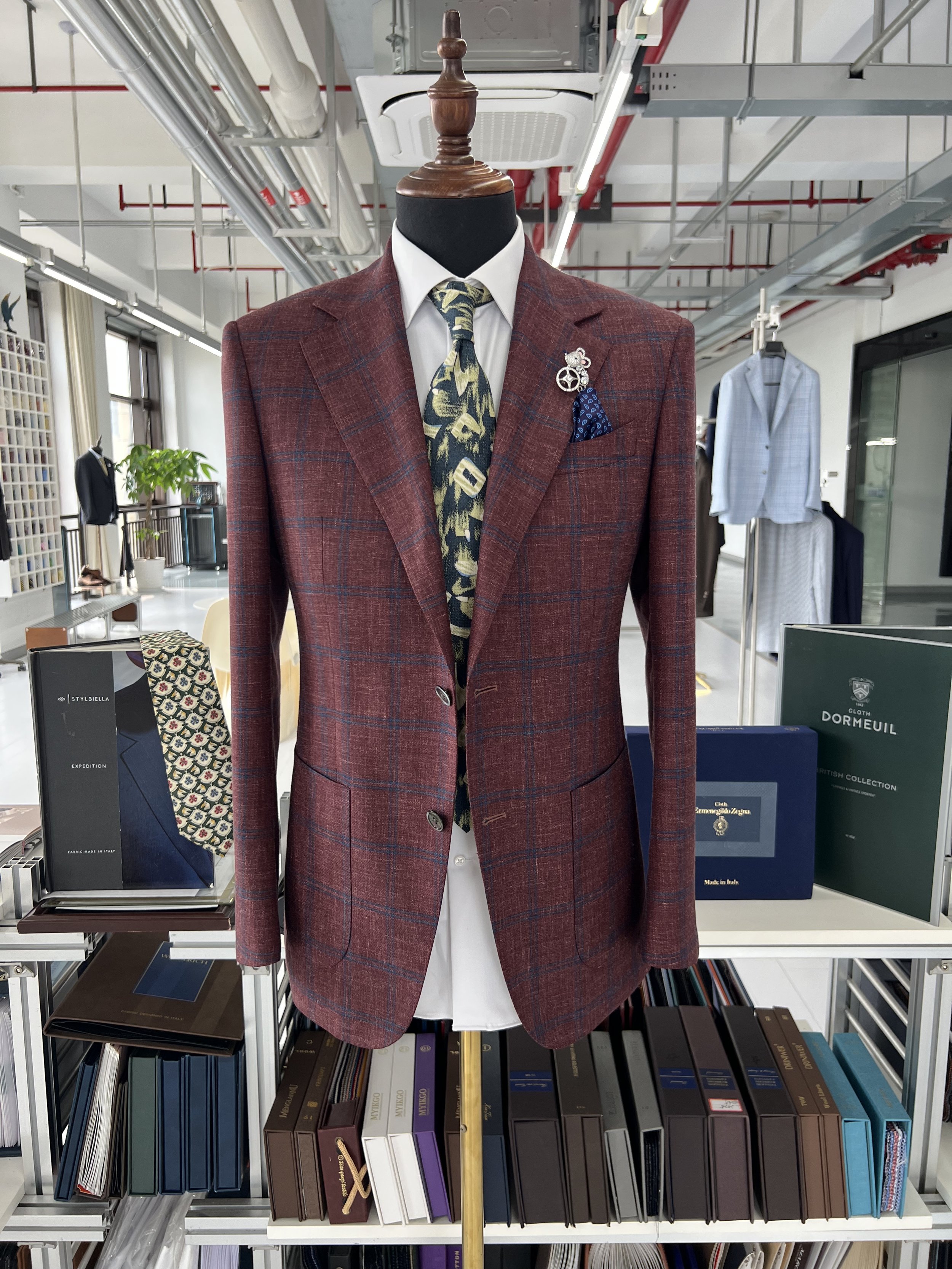 Display of a men's suit on a mannequin in a store. The suit is burgundy with a plaid pattern, worn with a white shirt, a patterned tie, and a blue pocket square. The mannequin has decorative pins on the lapel.