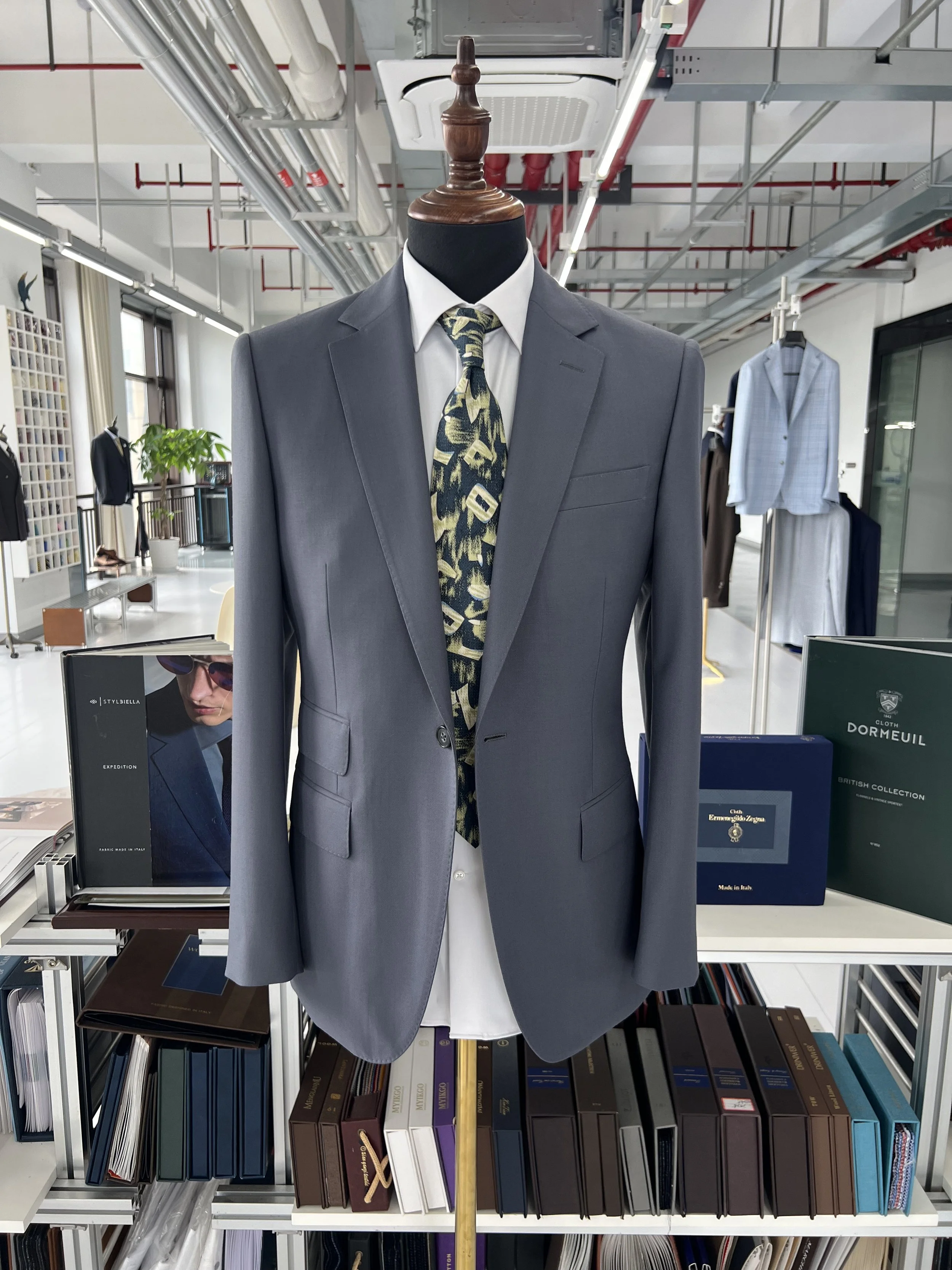 Mannequin wearing a gray suit jacket, white collared shirt, and a patterned tie, with books and office supplies on shelves in the background.