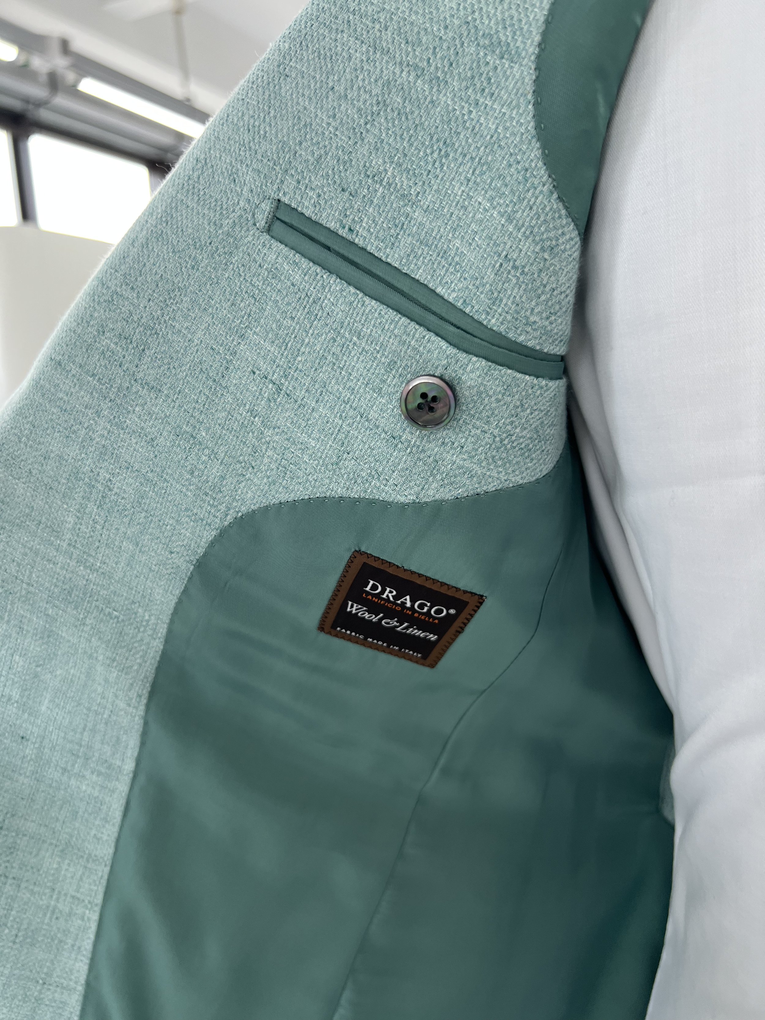 Close-up of the interior of a light green jacket, showing a zippered pocket, a button, and a brand label reading 'DRAGO Wool & Linen'.