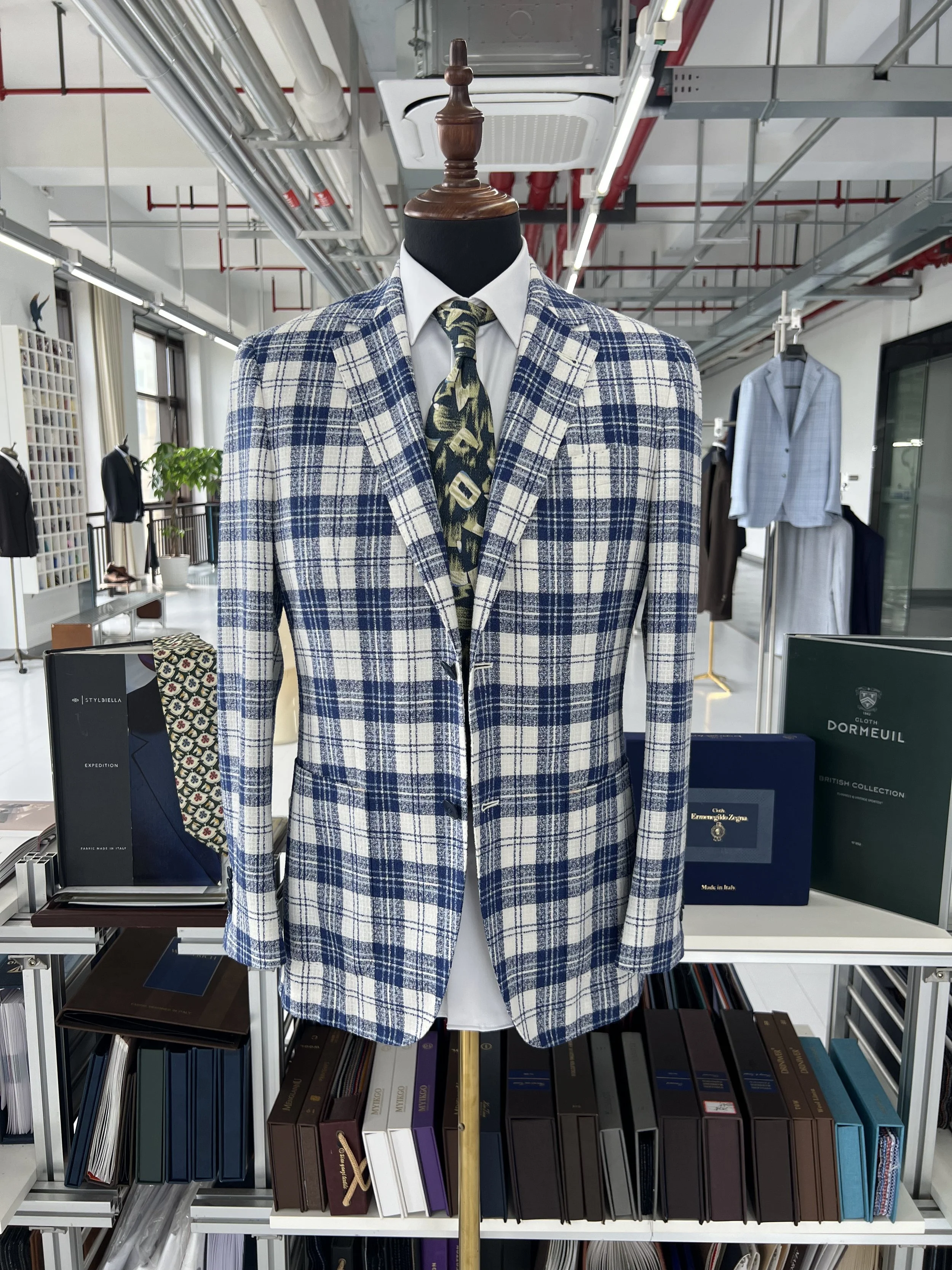 A mannequin dressed in a blue and white plaid blazer, a white shirt, and a patterned tie, displayed in a store with books and clothing in the background.