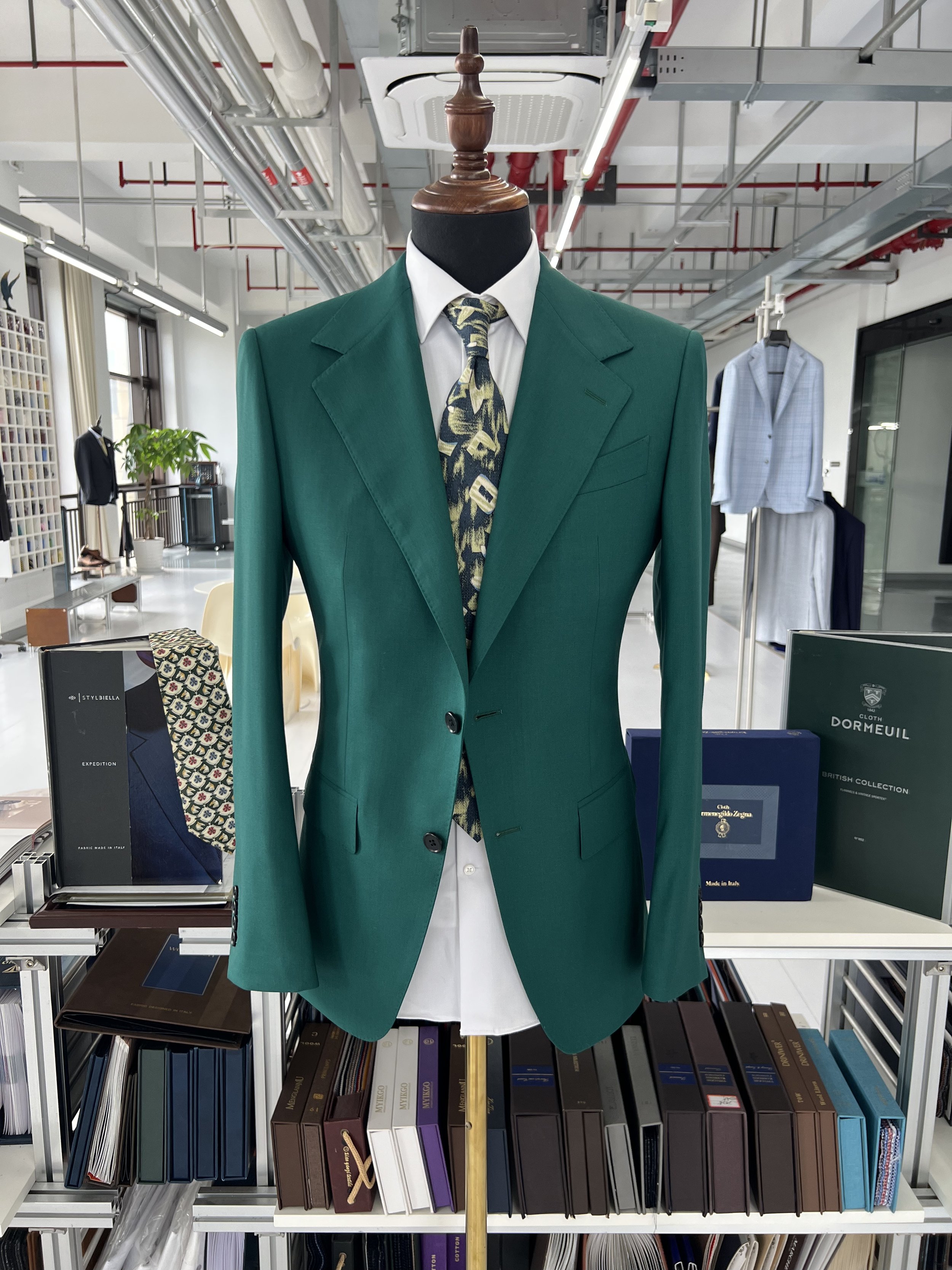 A mannequin dressed in a green suit jacket, white shirt, and a patterned tie, displayed in a store with shelves of fabric and books in the background.