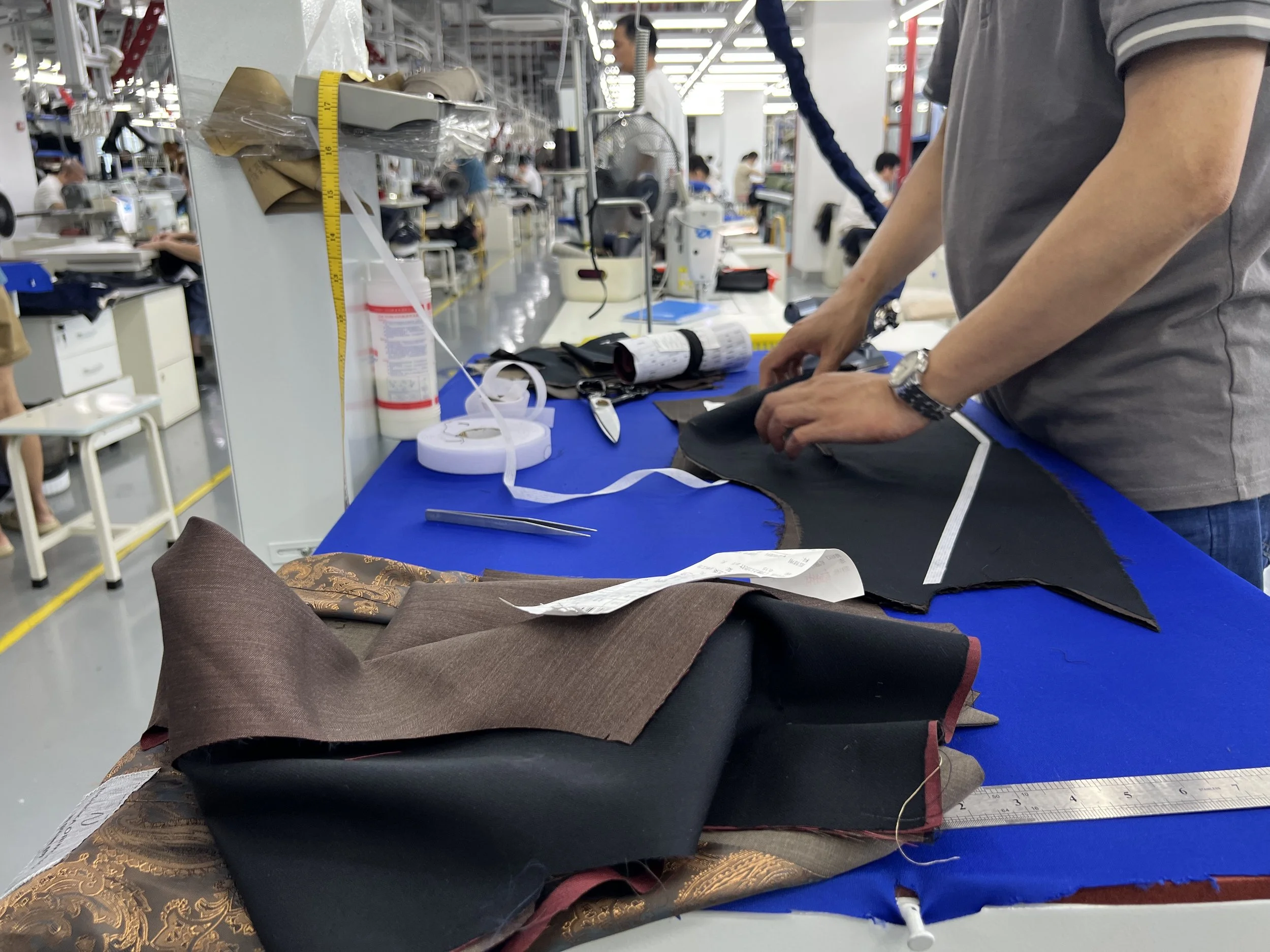 A person working on fabric pieces at a workstation in a factory or workshop, surrounded by sewing tools and materials.