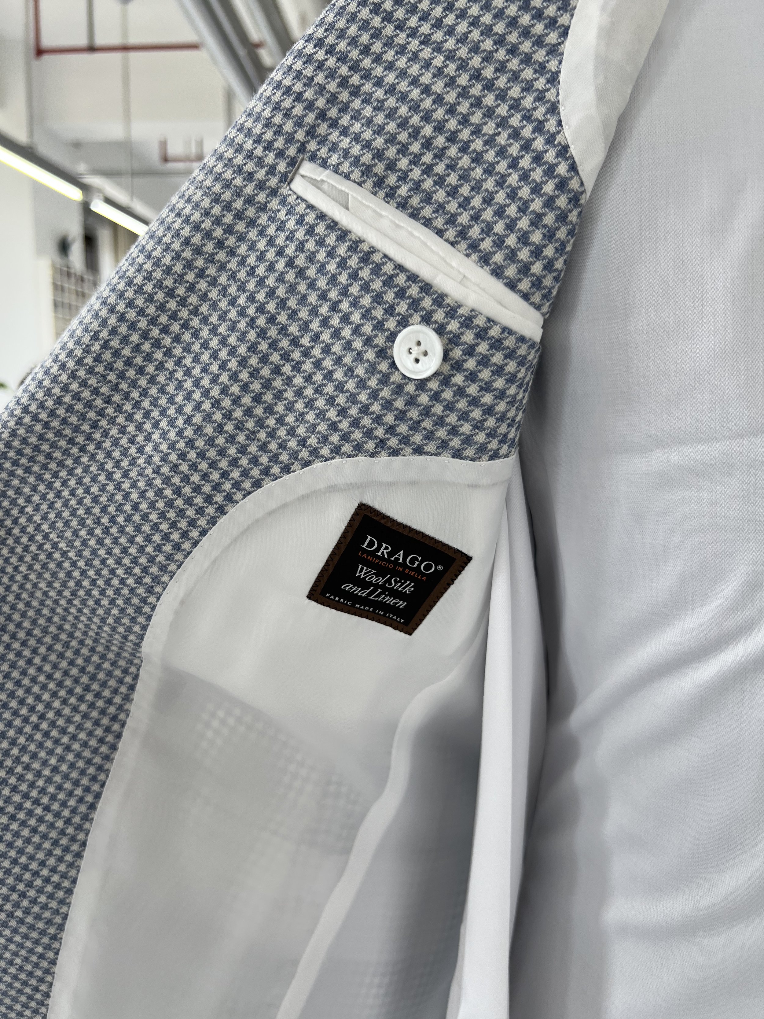 Close-up of a men's suit jacket with a houndstooth pattern, white lining, a white button, and a label reading 'DRAGO Lanificio in BIELLA Wool Silk and Linen Fabric Made in Italy'.