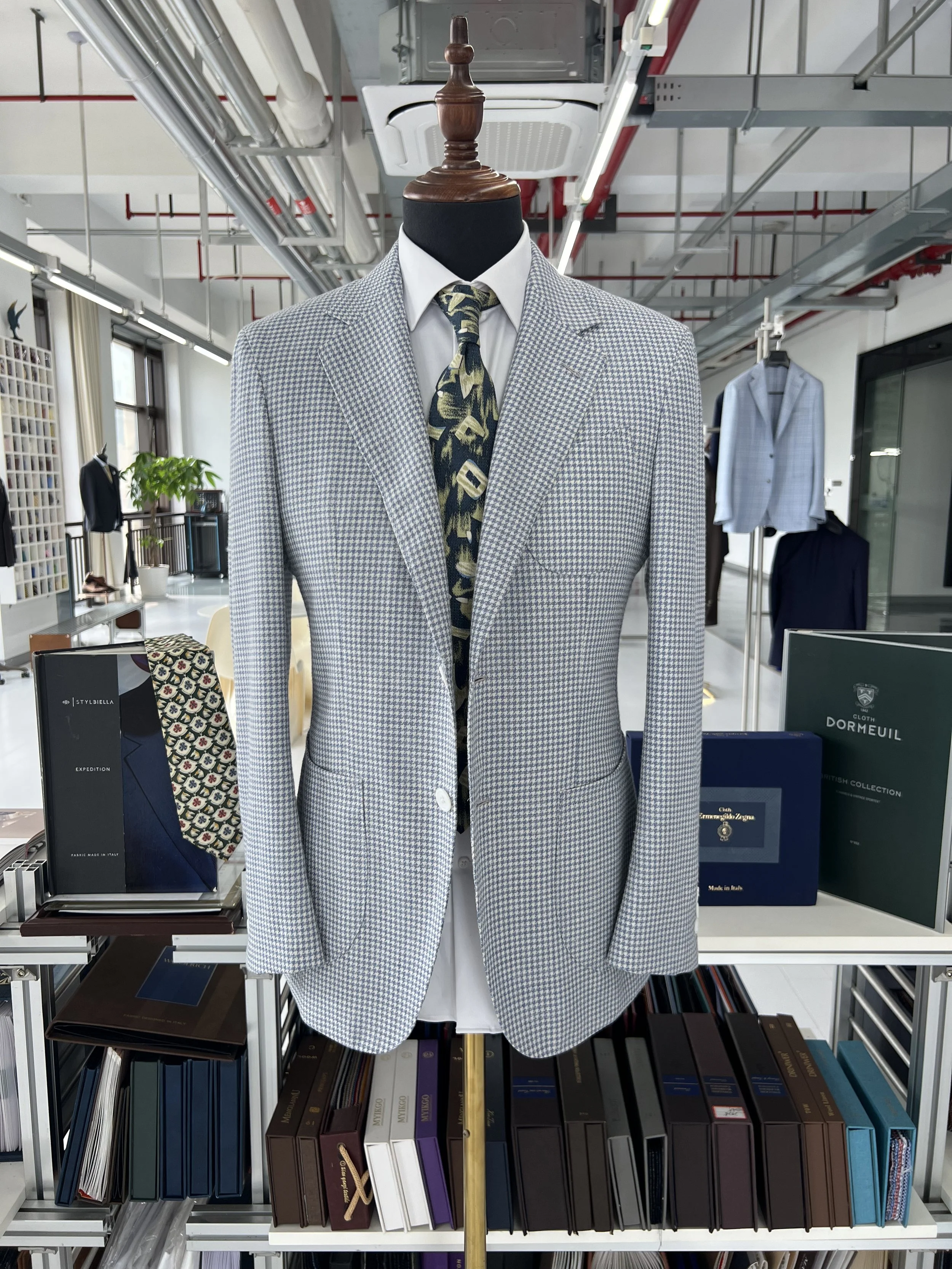 A mannequin dressed in a light-colored, checkered suit with a white shirt and a patterned tie, displayed in a retail store with shelves of books and other suits in the background.