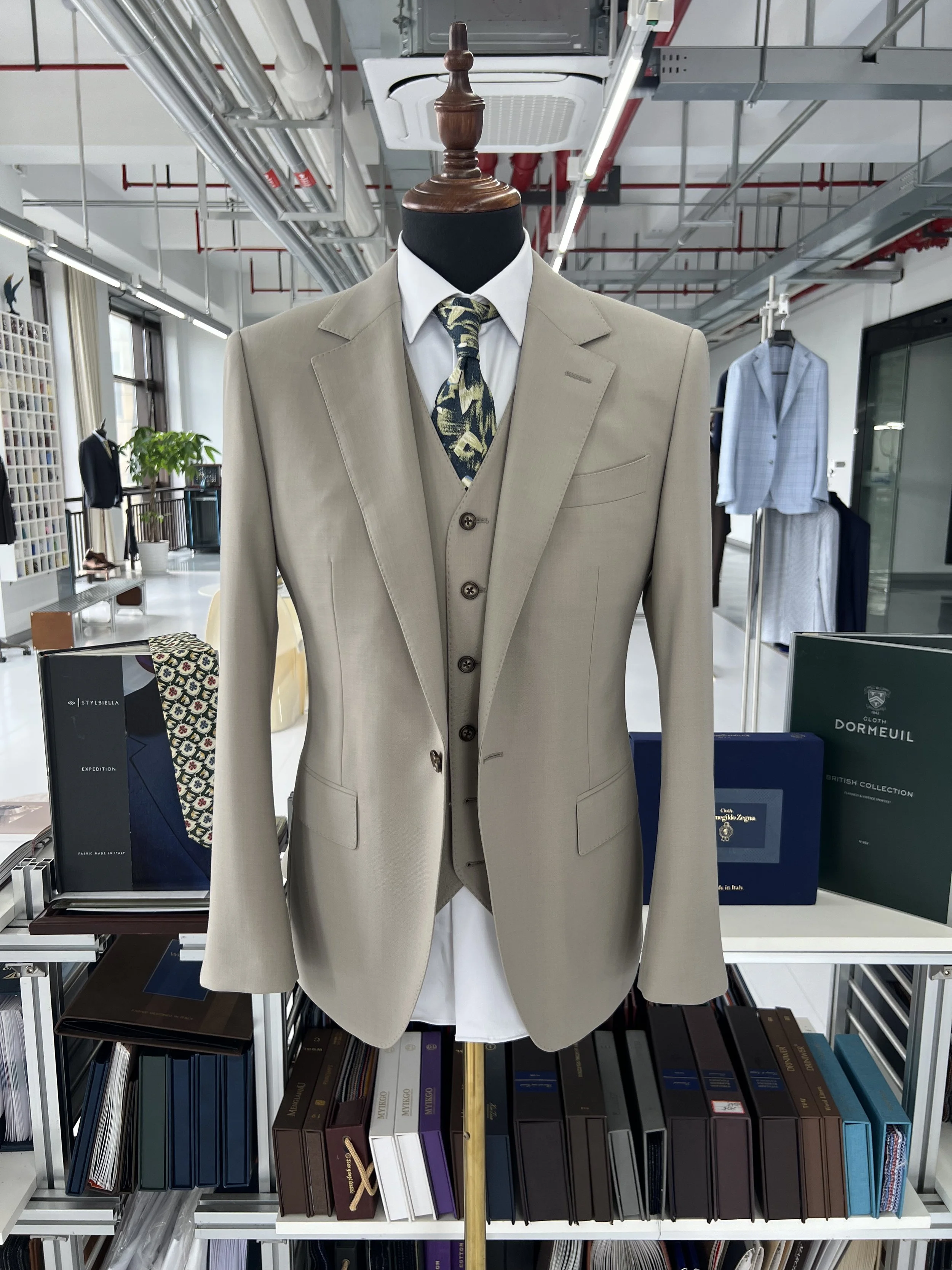 A mannequin dressed in a beige suit with a white shirt and a patterned tie, displayed in a store. The store has shelves with folders, books, and clothing items visible in the background.
