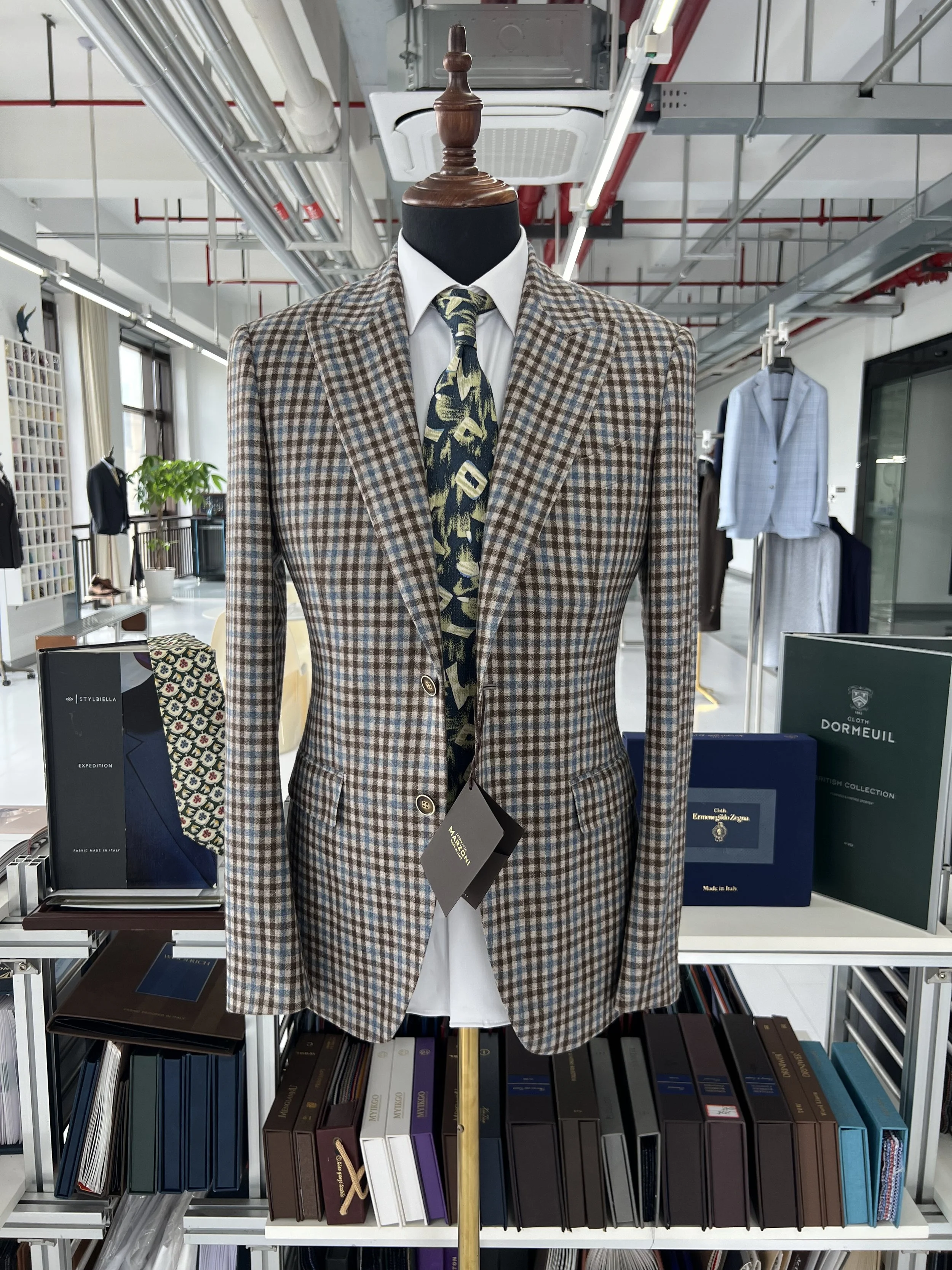 A mannequin dressed in a plaid brown and beige suit jacket, white dress shirt, and a patterned tie, displayed in a retail store with shelves of books and clothing in the background.