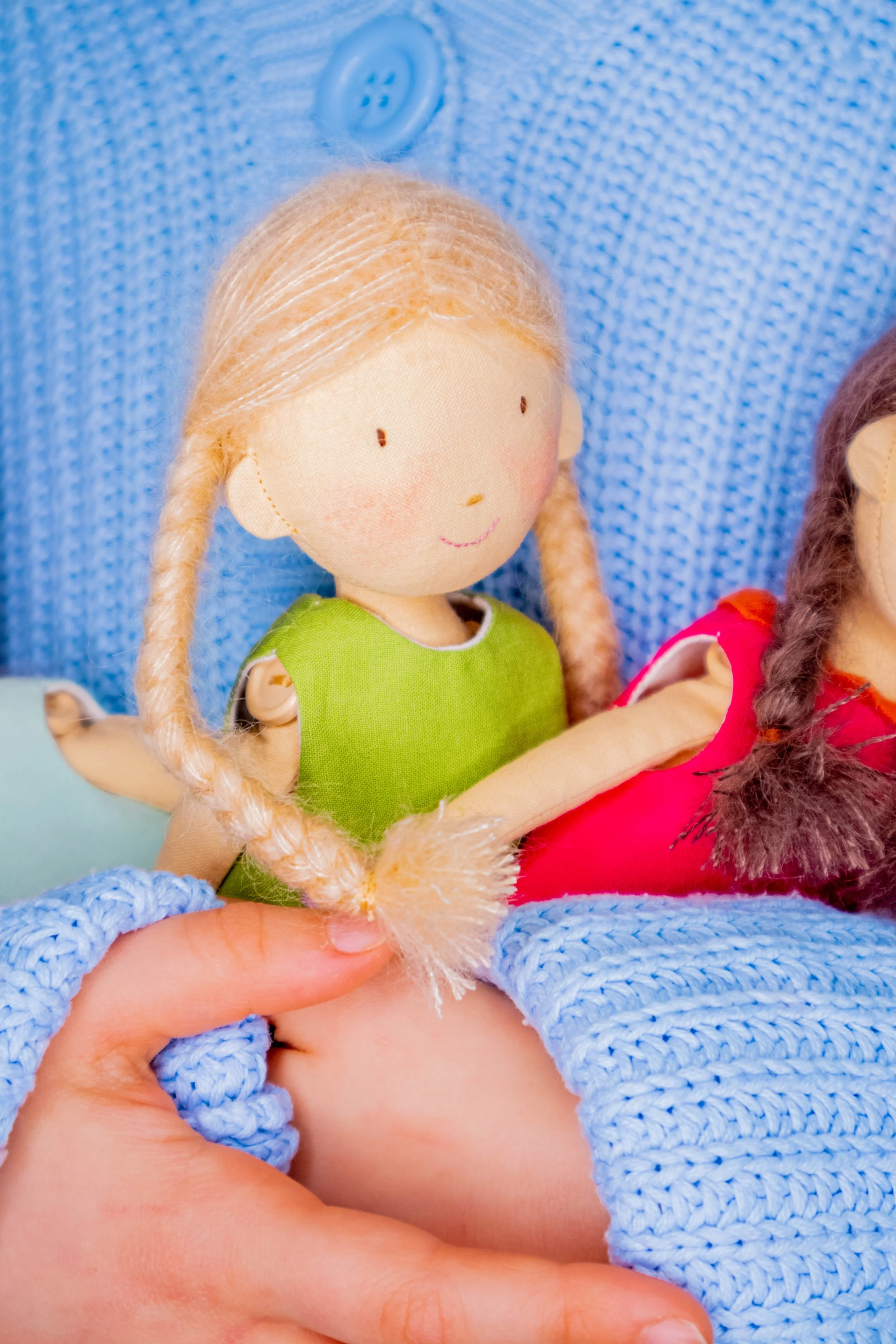 Close-up of a doll with blonde hair, wearing a green dress, held by a hand with blue knitted sleeve. Partially visible is another doll with brown hair in red clothing. They are in front of a blue fabric background.