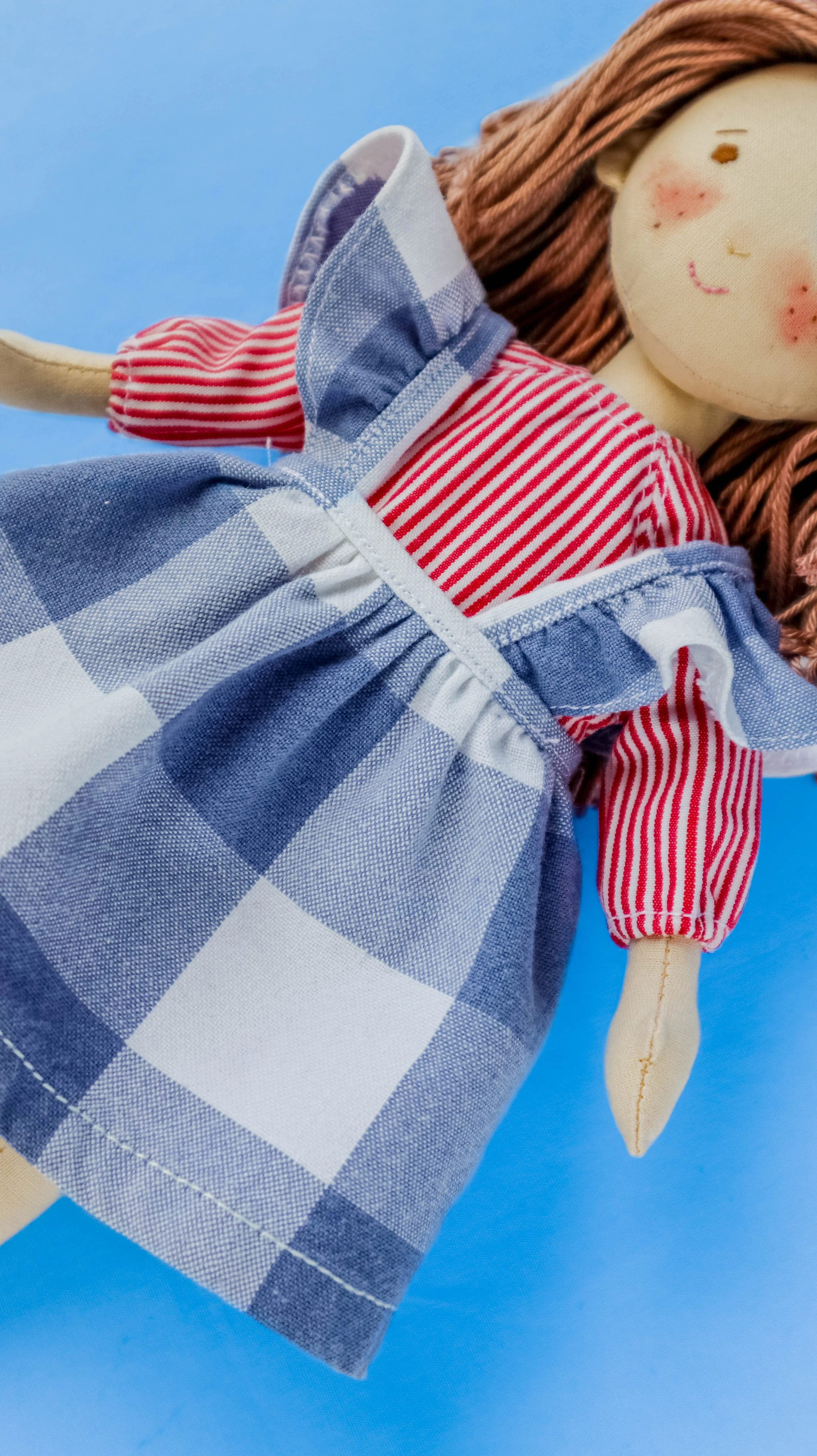 Close-up of a handmade doll with a smiling face, rosy cheeks, wearing a red and white striped shirt, a blue and white checkered apron, and a pinkish-brown braided hairstyle, set against a blue background.