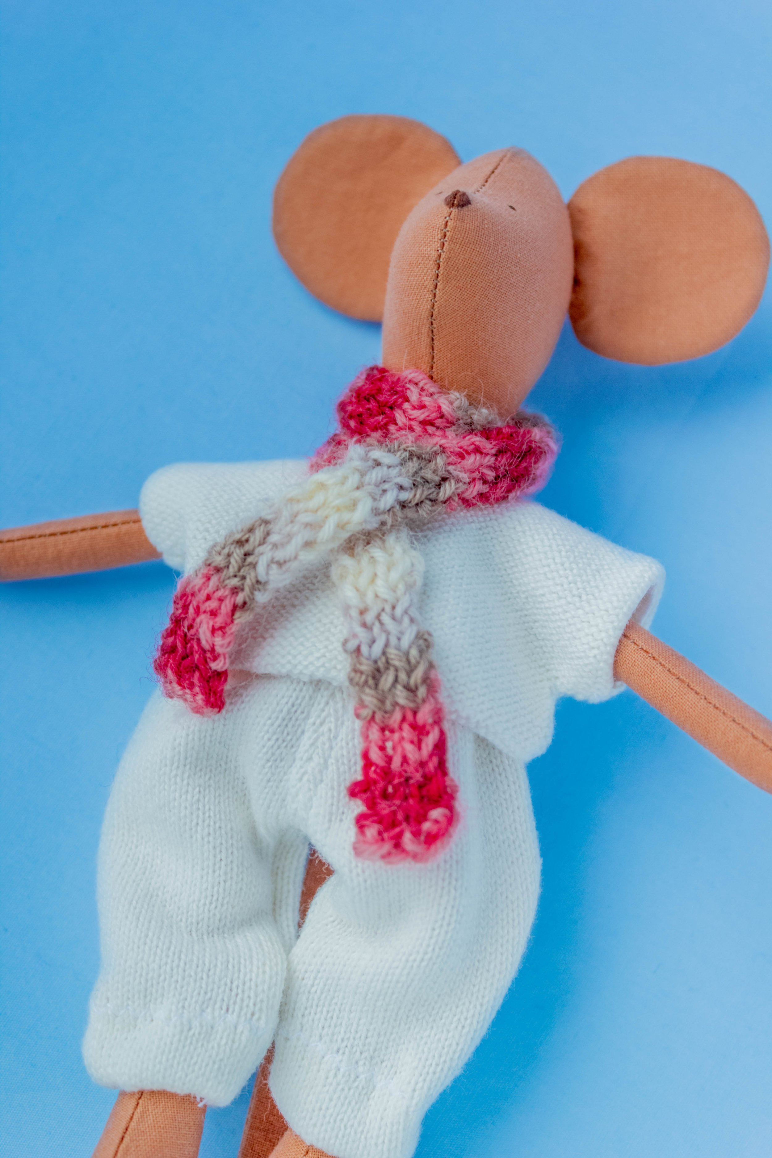 A stuffed toy mouse wearing a white dress and a multicolored knitted scarf, set against a blue background.
