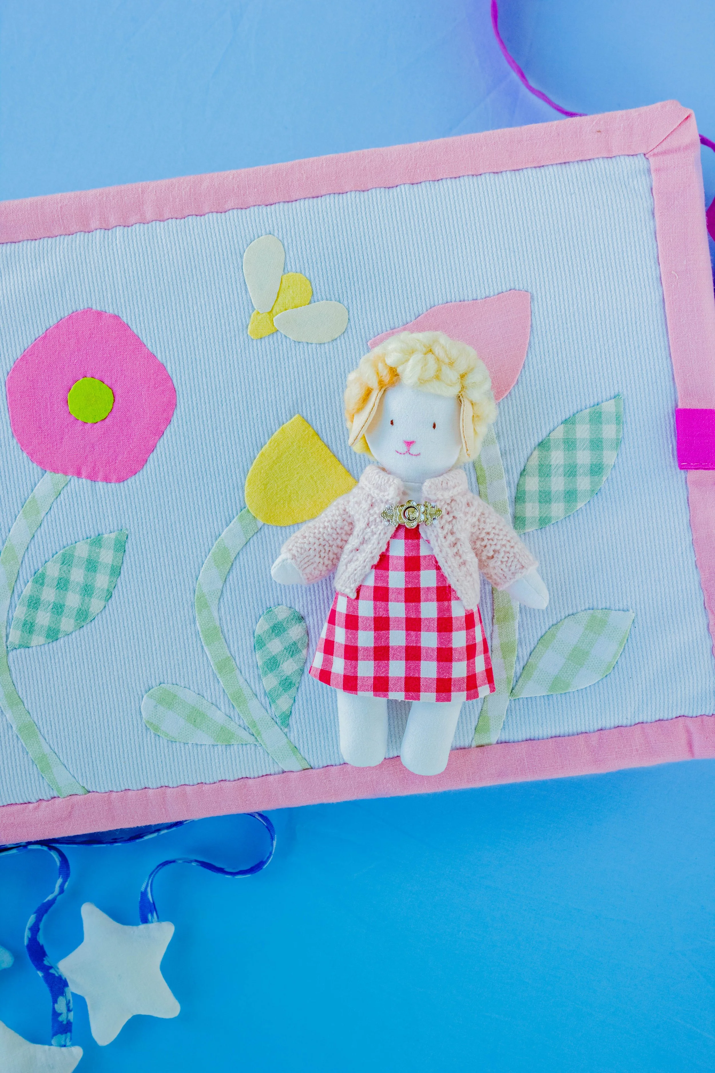 A fabric wall hanging with a plush doll of a lamb in a pink and red checkered dress, surrounded by felt flowers, leaves, a butterfly, and stars on a blue background.