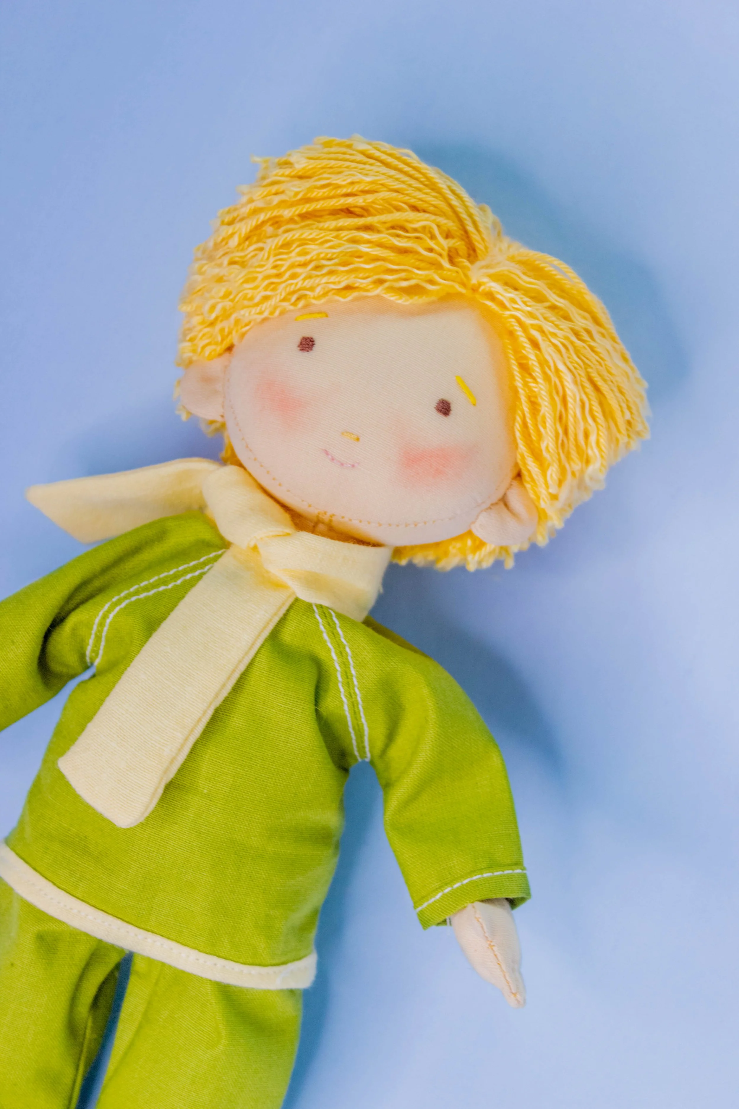 A plush doll with yellow yarn hair, wearing a green dress with white trim, against a light blue background.